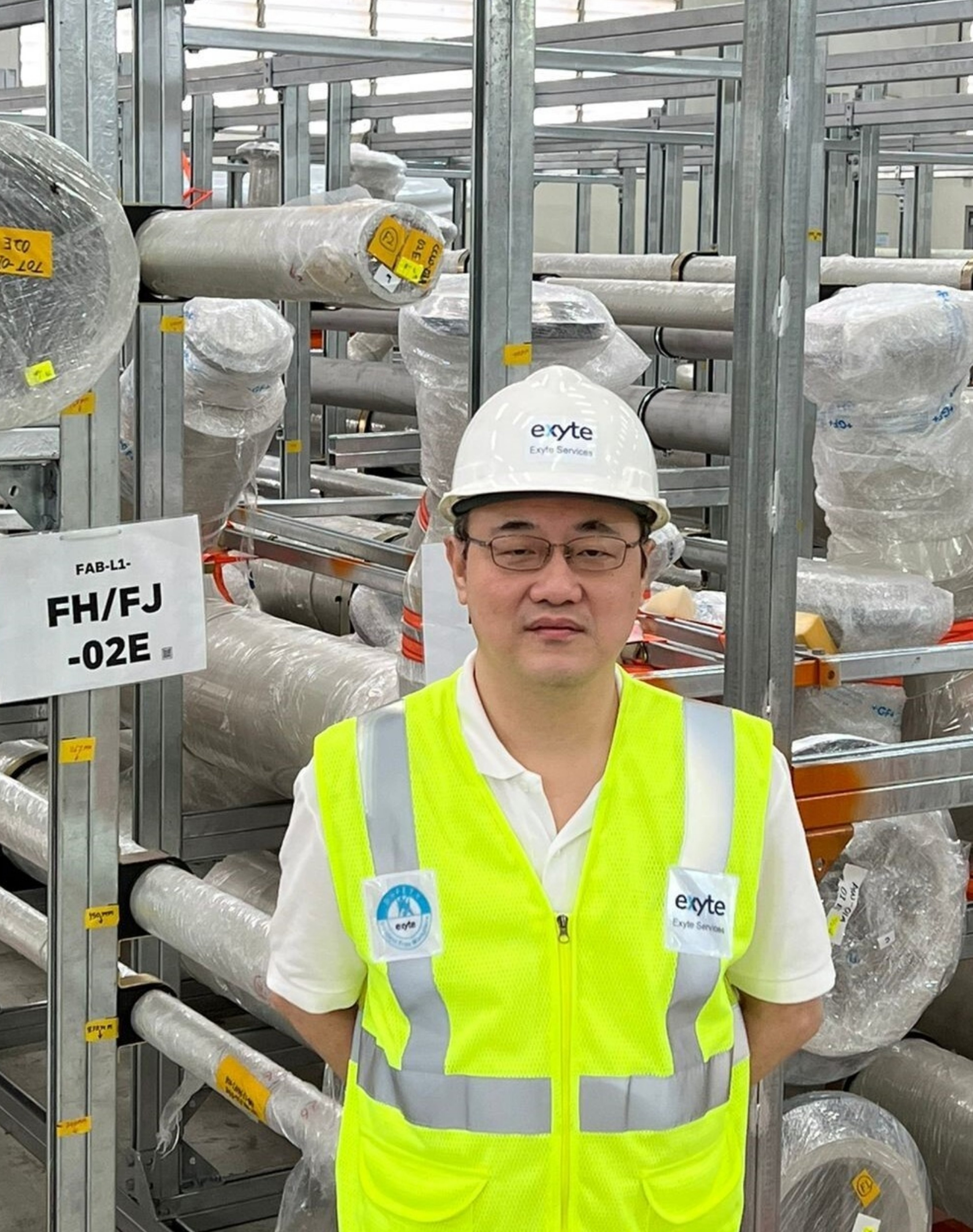 A portrait of Ng Aik Beng. He is wearing a white hard hat and a high-visibility yellow safety vest with the Exyte logo. He stands in an industrial environment, surrounded by wrapped materials and pipes.