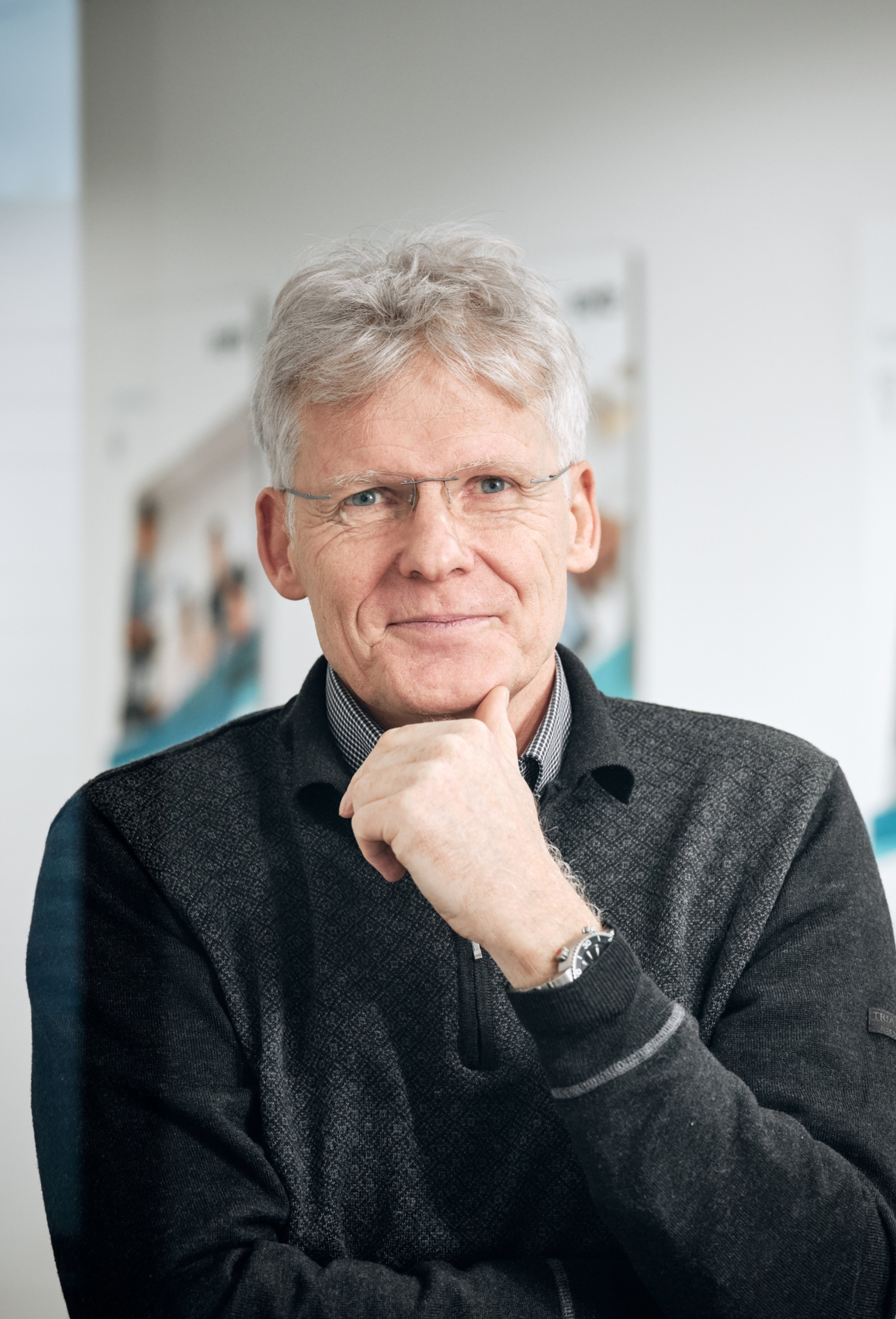 Manfred Engelhard, standing with a thoughtful expression. He is wearing glasses and a dark sweater, with his hand resting near his chin.