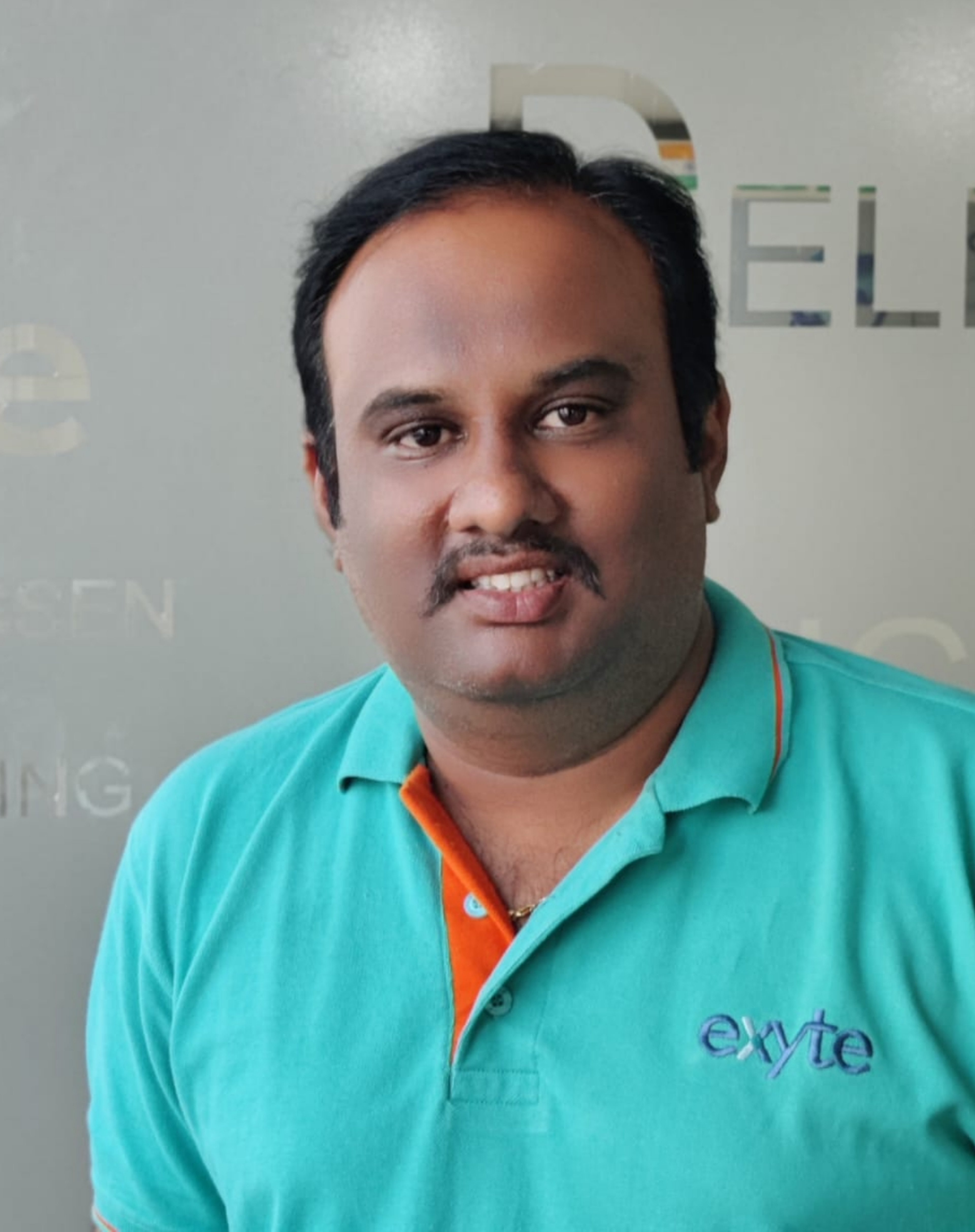 A portrait of Saravanan Natarajan. He is wearing a turquoise Exyte polo shirt and smiling gently at the camera.