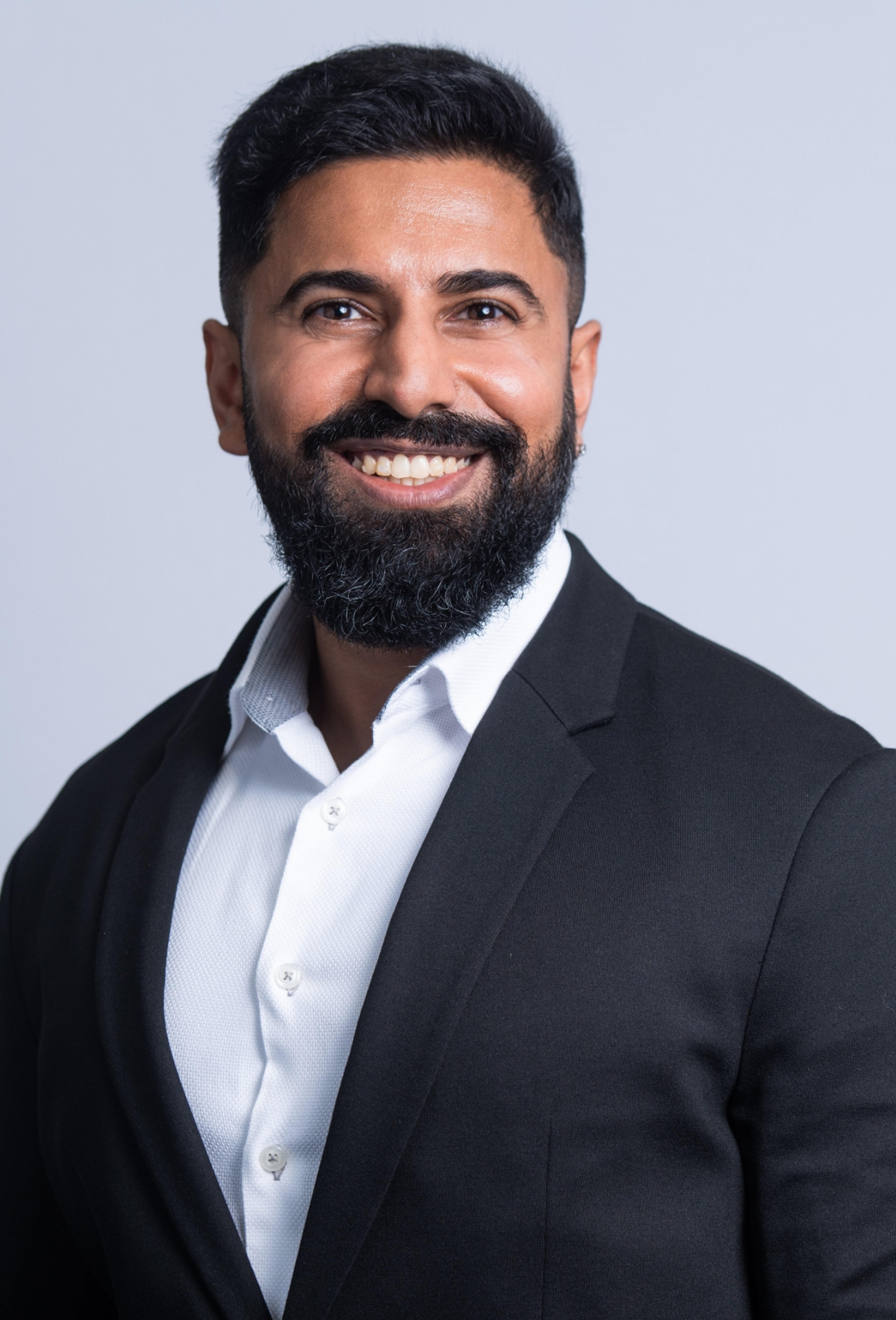 Taib Shabbir, smiling and dressed in formal business attire, consisting of a black suit jacket and white collared shirt. He has a full beard and short hair.