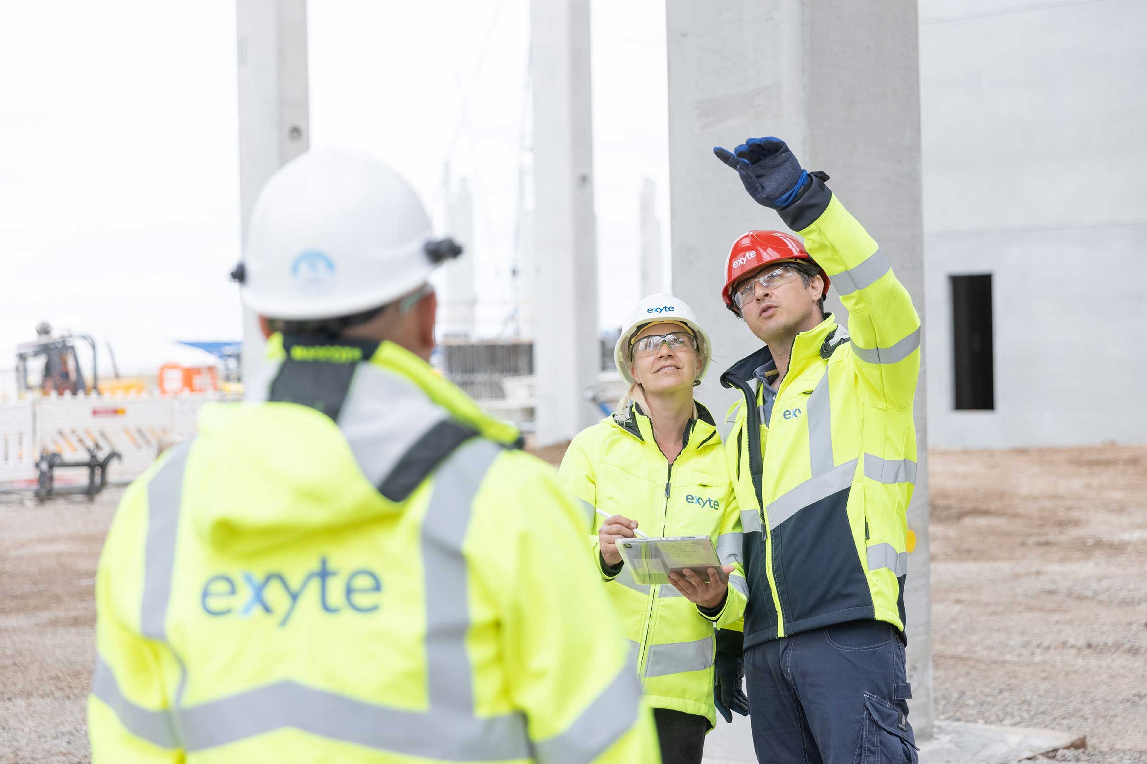 Three construction workers from Exyte, wearing high-visibility jackets and safety helmets, are standing on a construction site. One worker is pointing ahead, while another is holding a clipboard, and the third has their back to the camera.