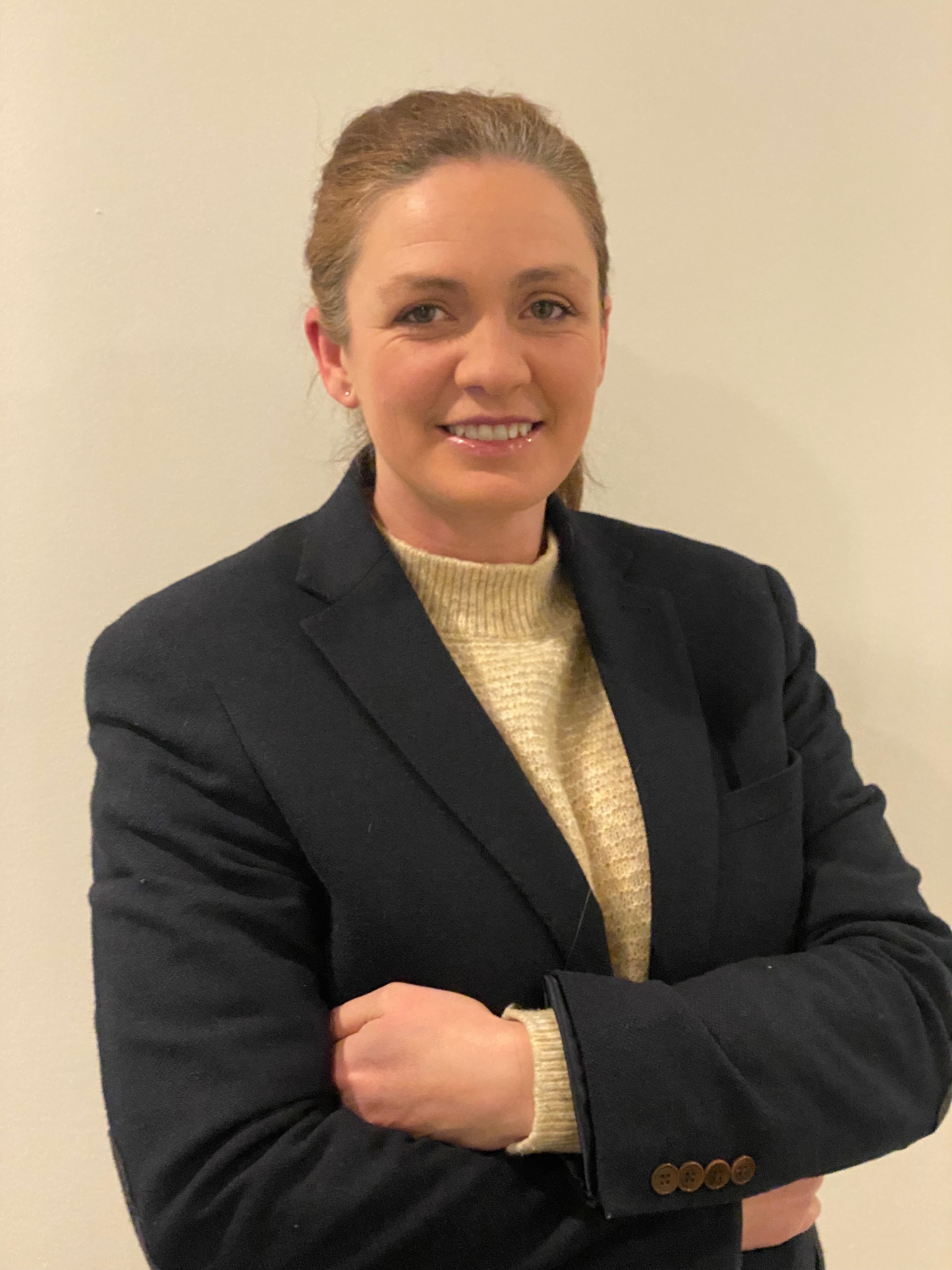 Liz Allan, smiling with arms crossed, wearing a dark blazer.