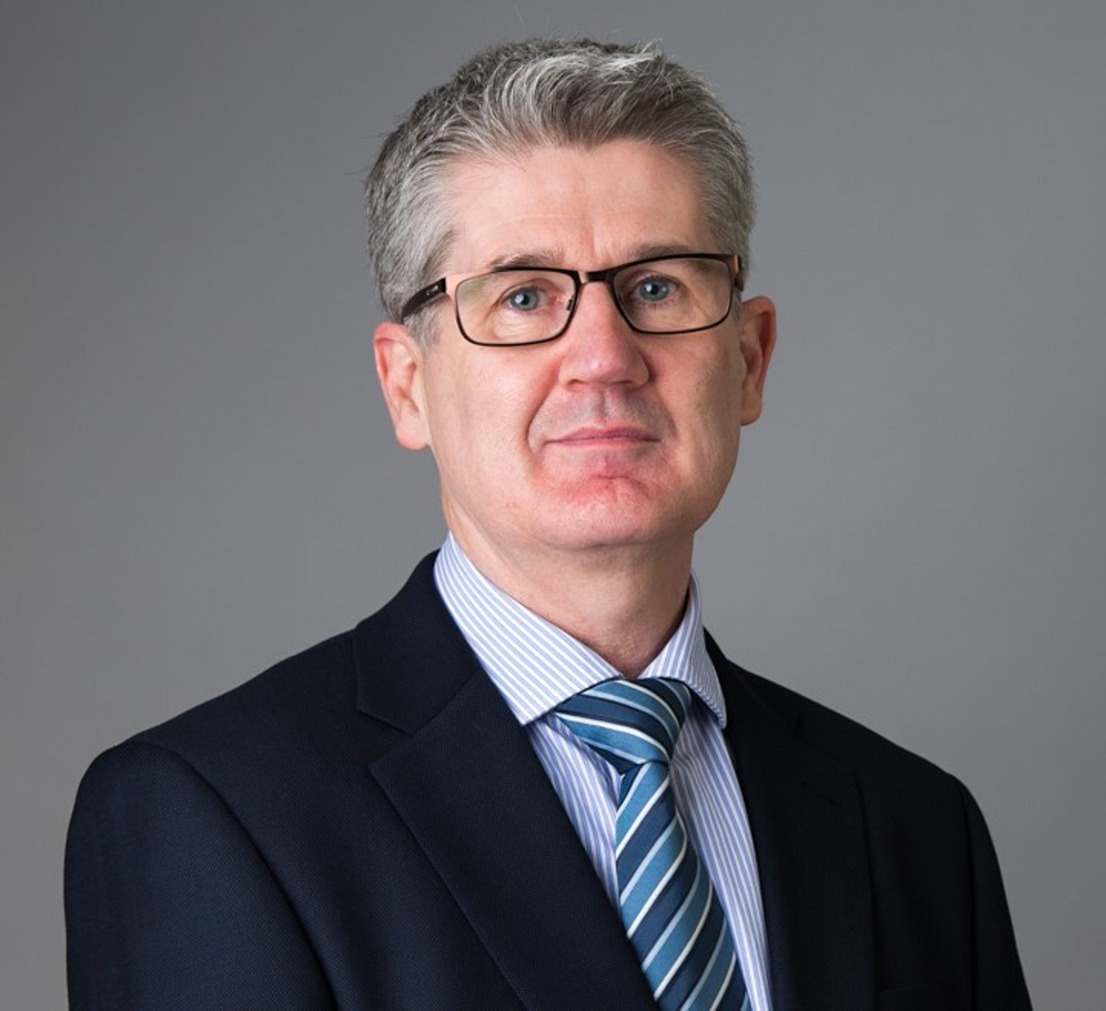 Padraic Brennan, wearing glasses and a formal suit with a blue striped tie, stands against a neutral background, giving a professional expression.
