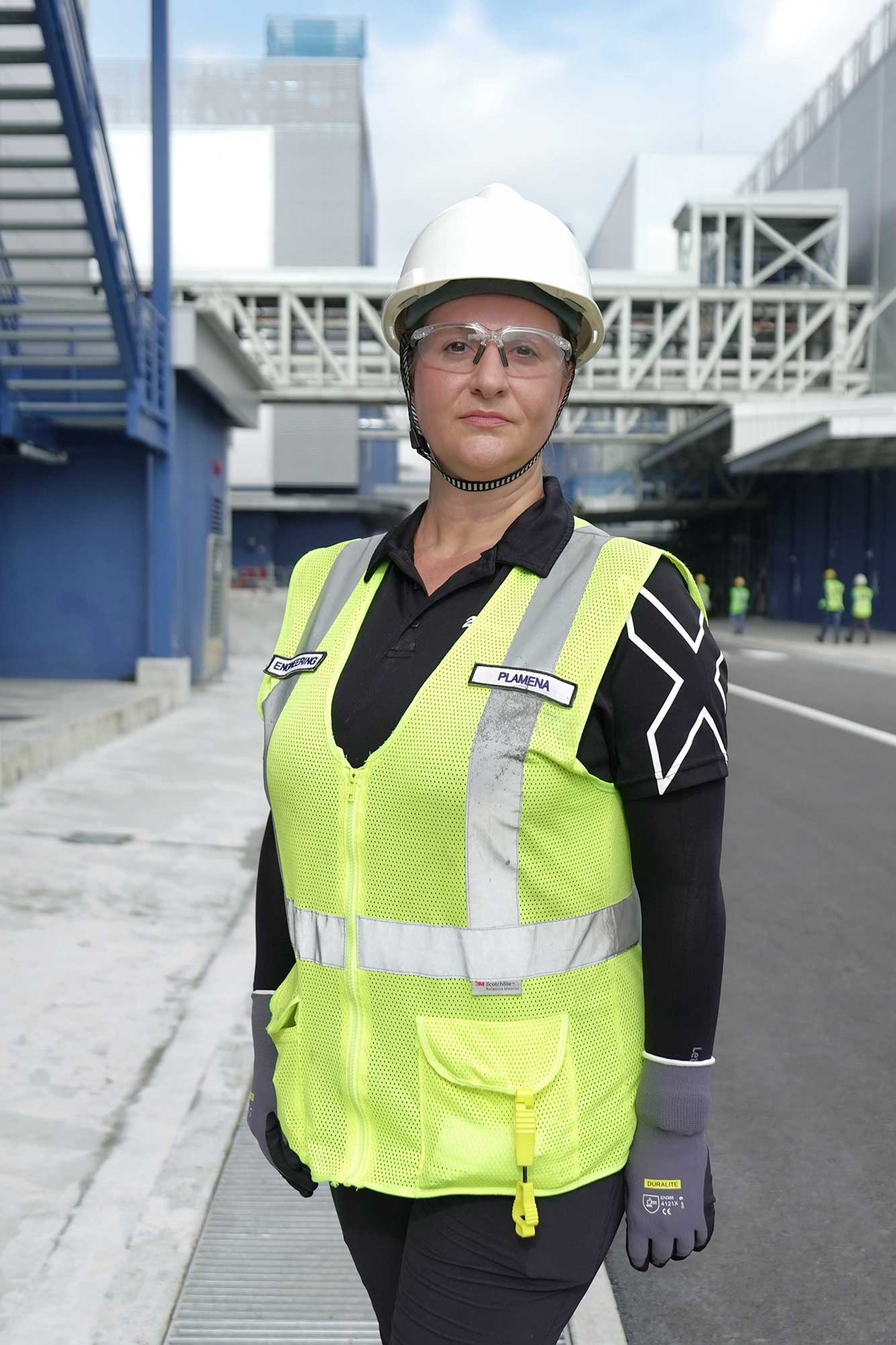 Plamena Petkova is shown at a construction site, wearing a white hard hat, safety glasses, and a reflective yellow vest with her name tag visible. She stands confidently in front of industrial structures, indicating her role in the engineering or construction field.