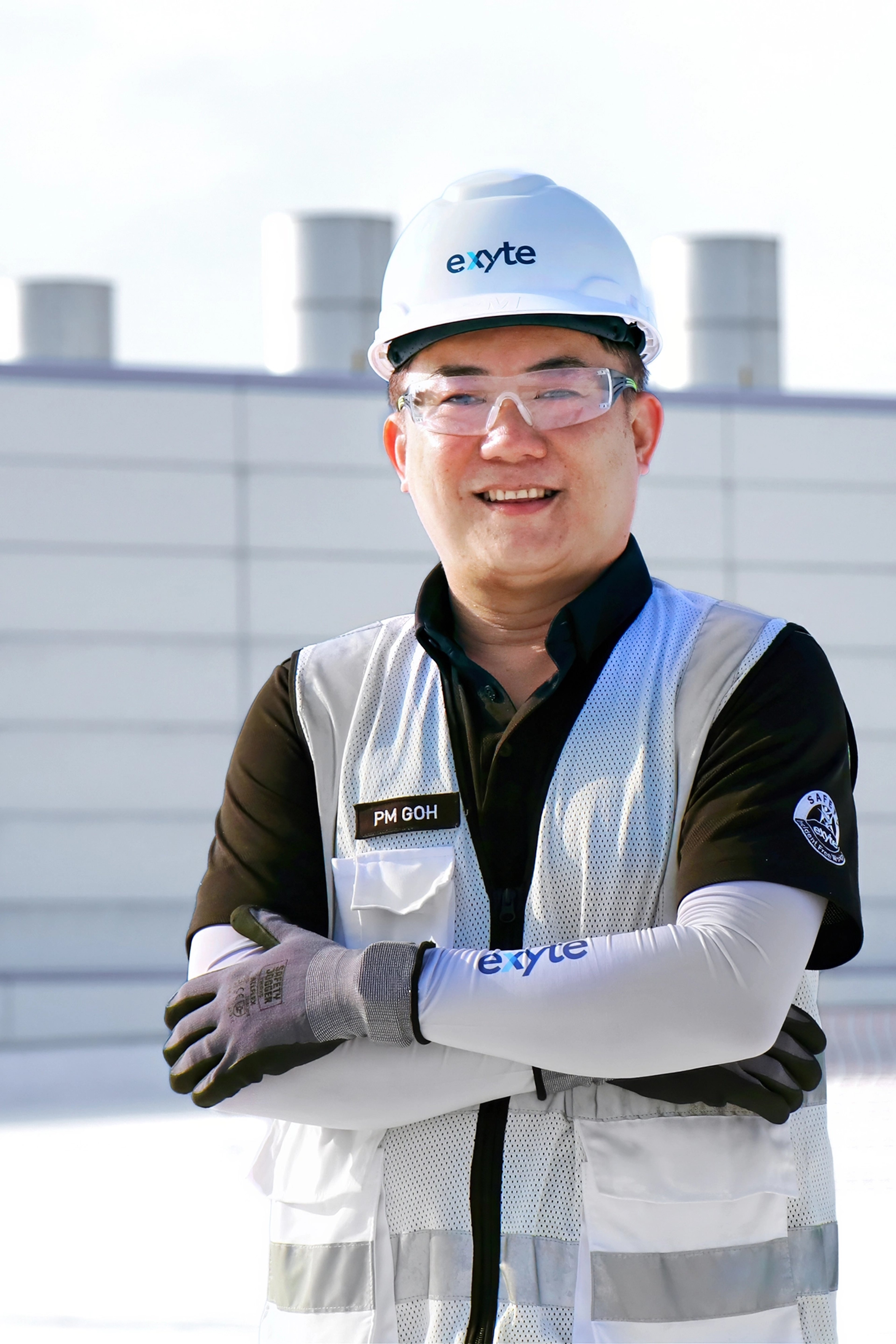 Puay Meng Goh, wearing safety gear including a hard hat, protective glasses, and gloves, stands confidently with his arms crossed, smiling during work at an outdoor industrial site.