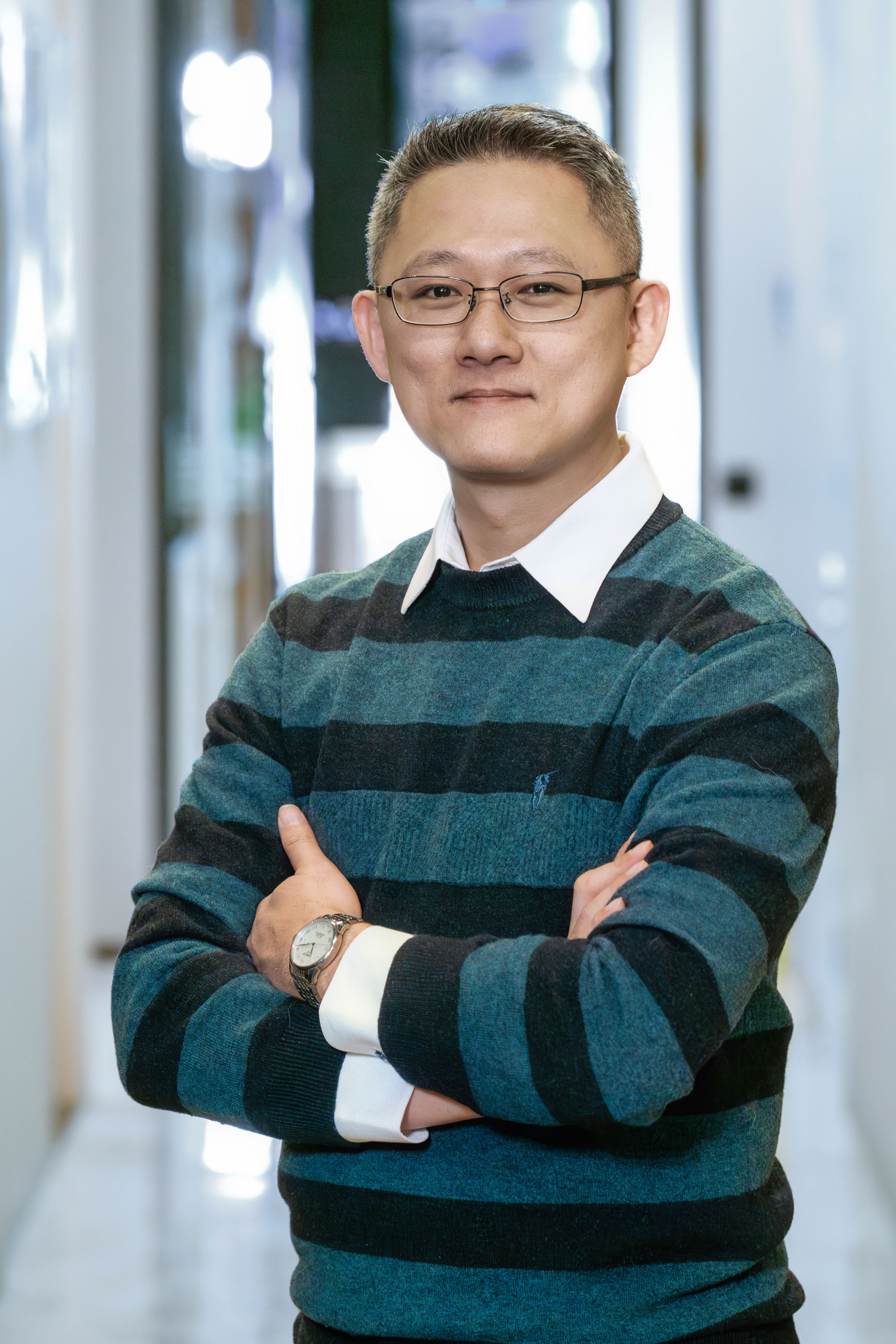 Tam Wing Hung stands confidently with his arms crossed, wearing glasses, a striped sweater, and a wristwatch. He smiles subtly while posing in a well-lit indoor setting.