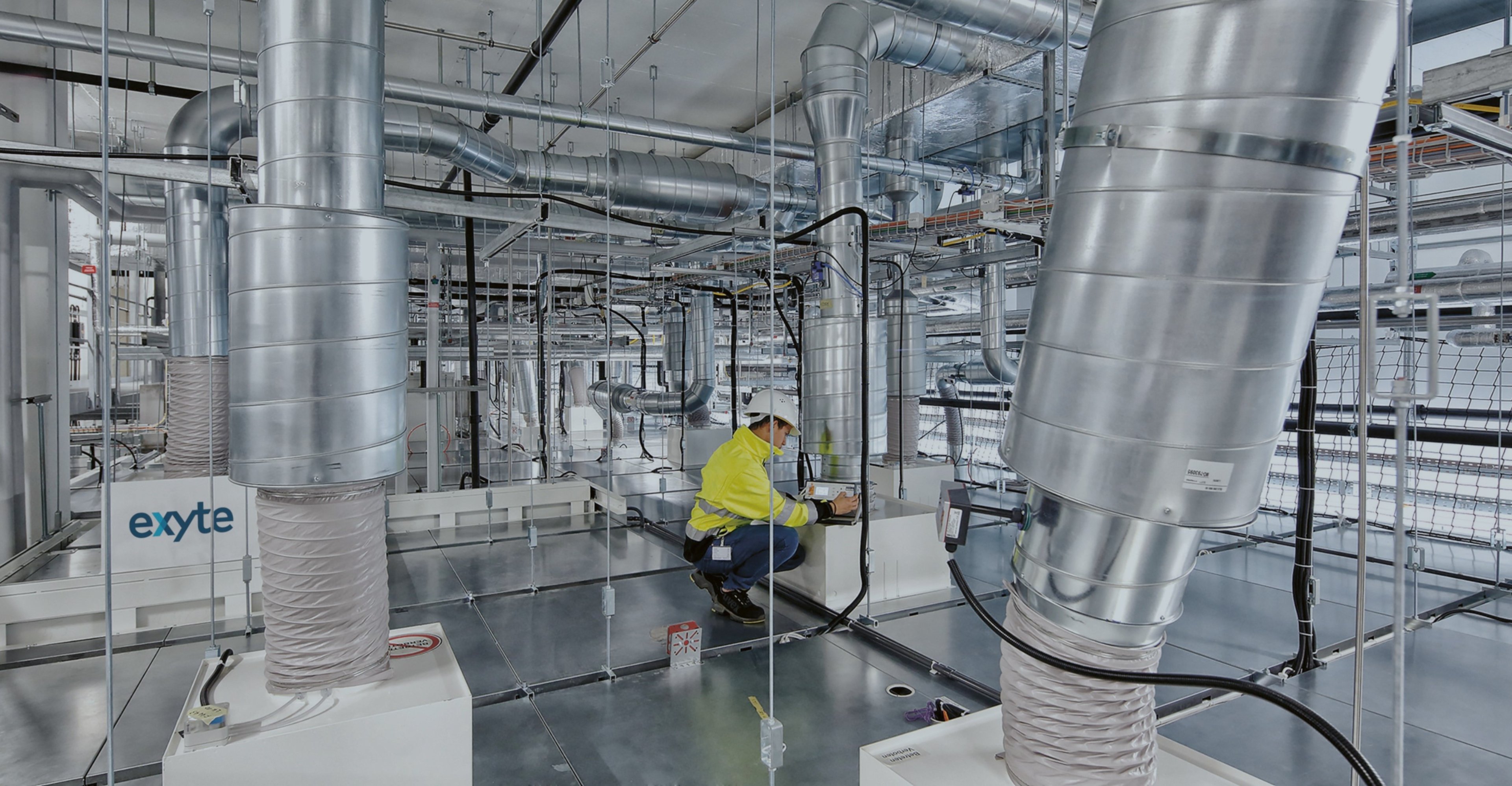 A technician working on advanced HVAC systems in a cleanroom environment with the Exyte logo visible, surrounded by complex ductwork and equipment, ensuring air quality and ventilation for precision manufacturing.