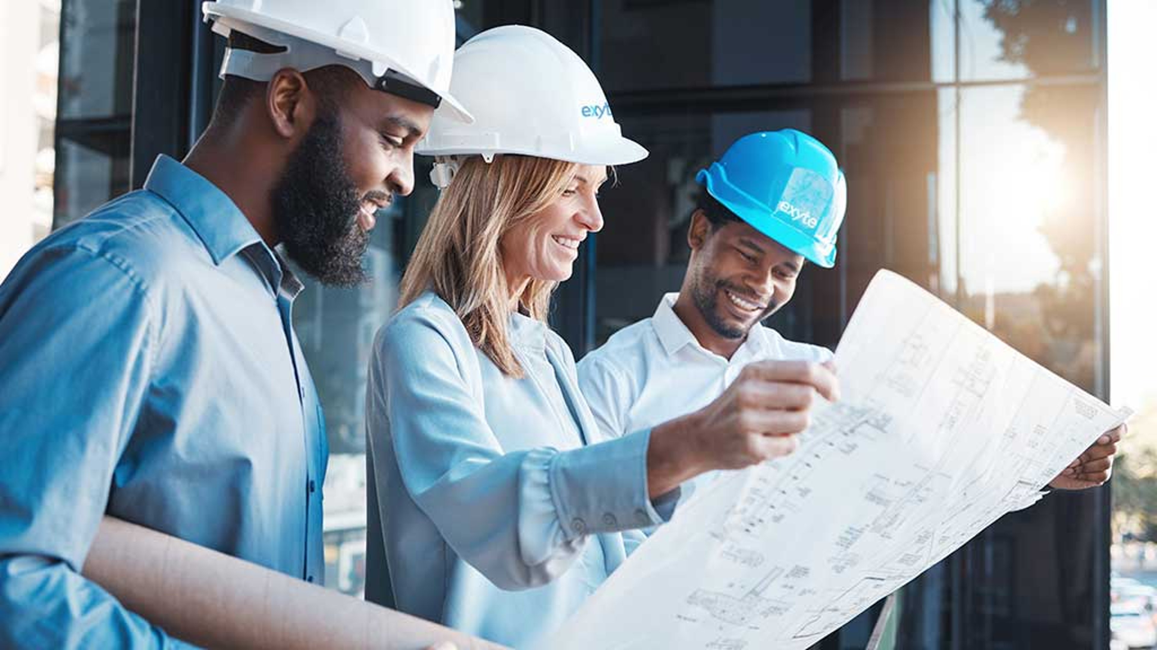 Three engineers in hardhats with the Exyte logo review a blueprint together outdoors, smiling and collaborating on the project.