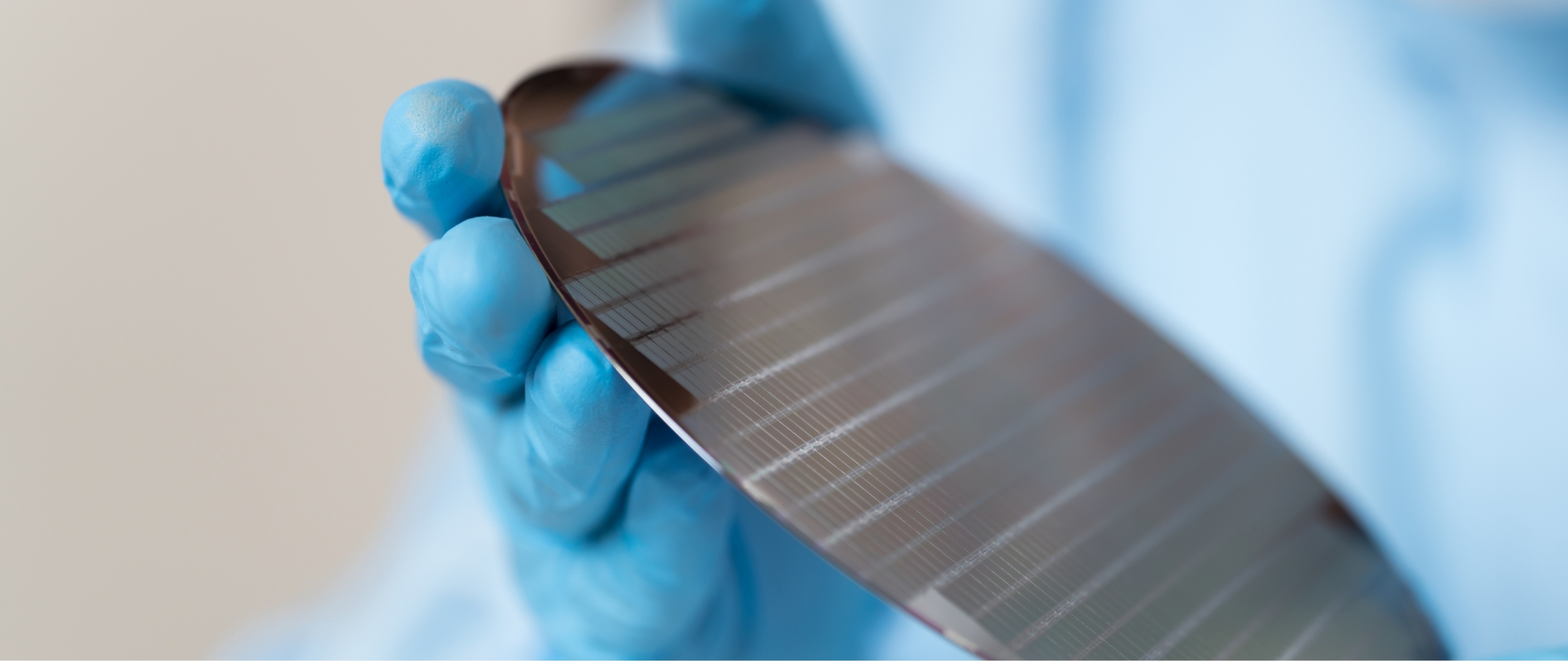 Gloved hand holding a semiconductor wafer, used in microchip and electronic component manufacturing, with precise lines visible on its reflective surface.
