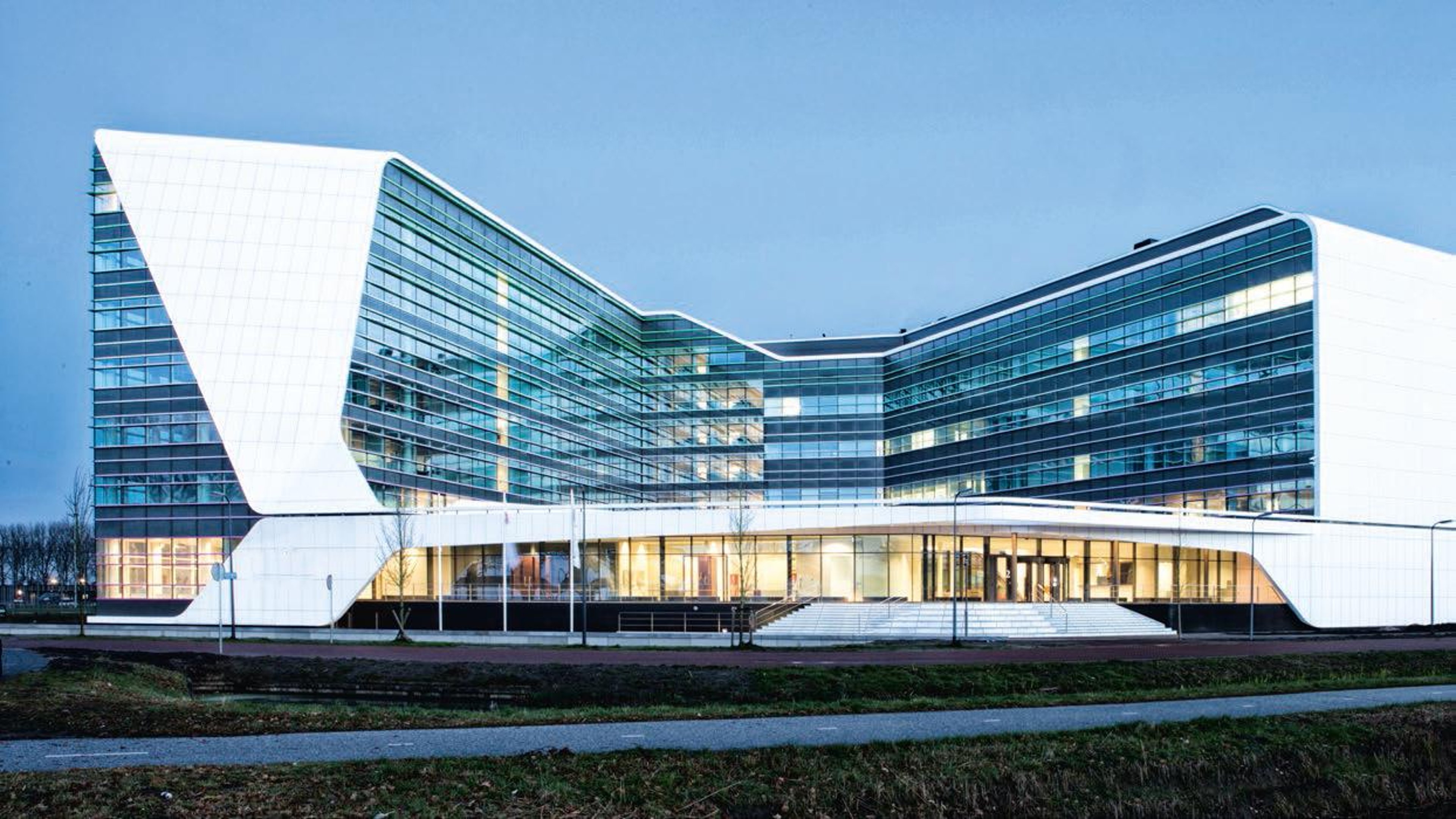 The image shows a modern building with a sleek, curved design featuring large glass windows and white exterior panels. The building appears to be an office or institutional facility, illuminated from within during dusk or early evening.