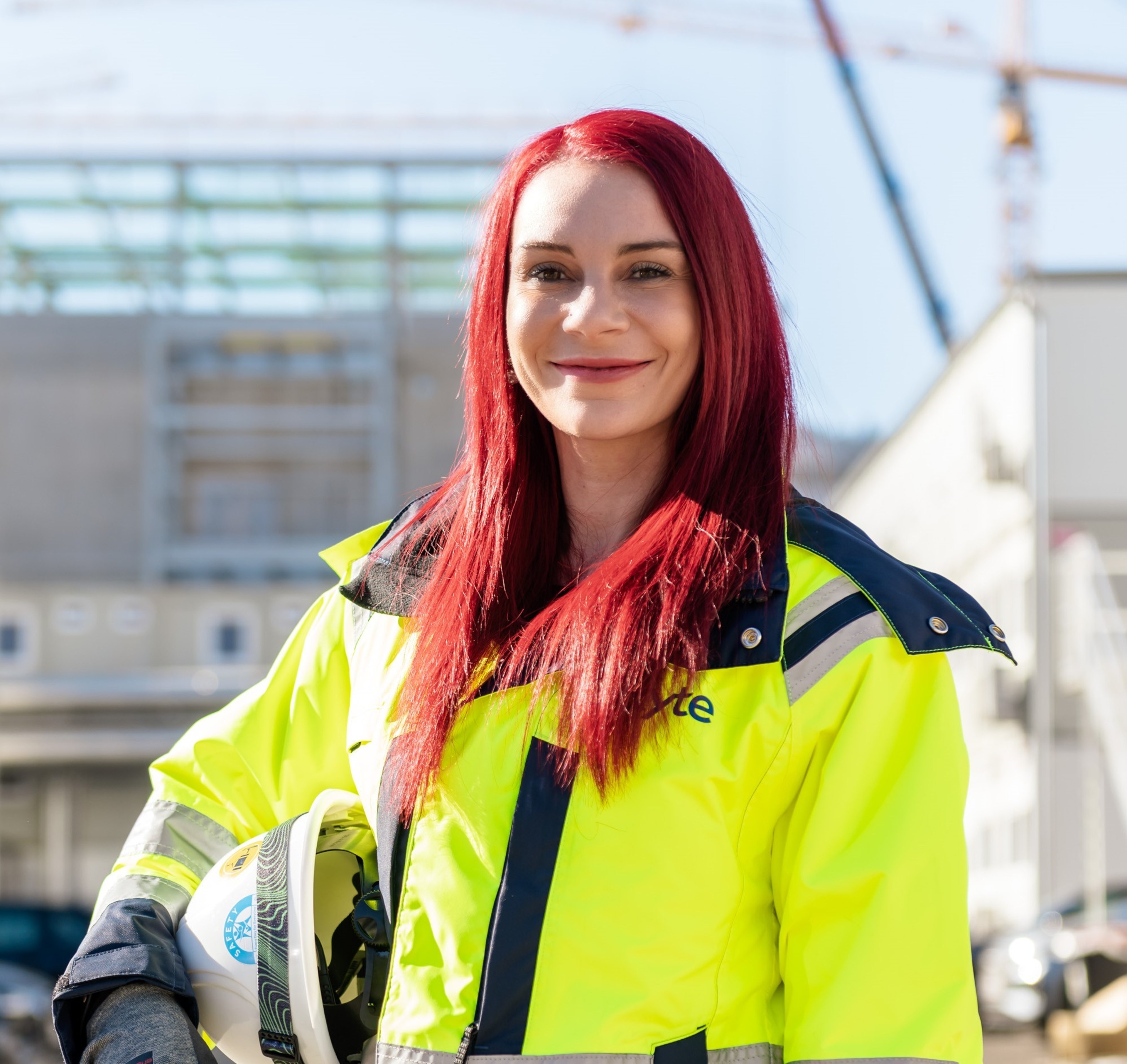 A portrait of Ciske Berrington, wearing a high-visibility construction jacket, smiling confidently at a construction site in the background. She holds a helmet in her hand, with her red hair standing out prominently.