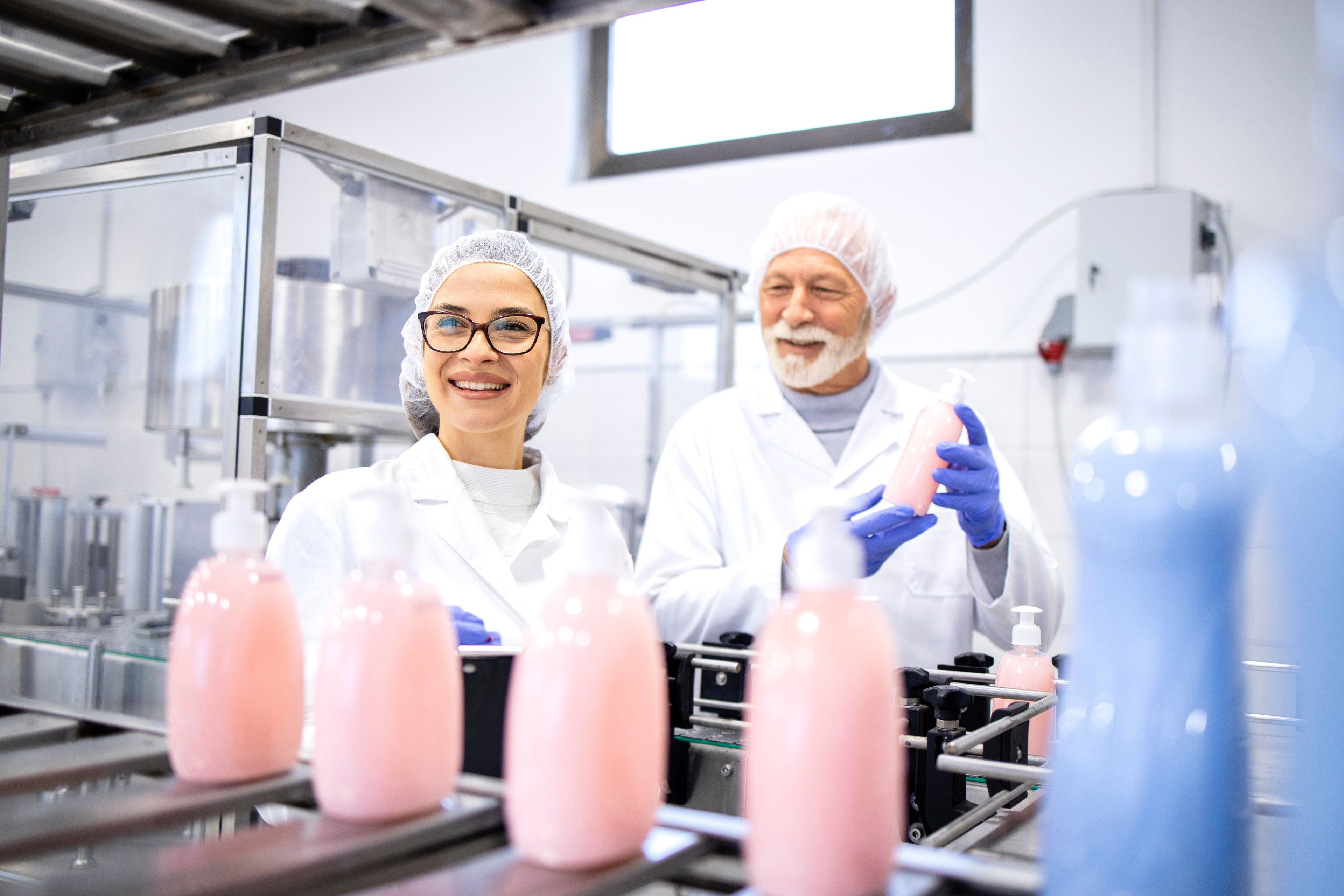 Two factory workers smiling as they work on a production line, inspecting bottles of liquid soap or shampoo in a clean manufacturing environment.