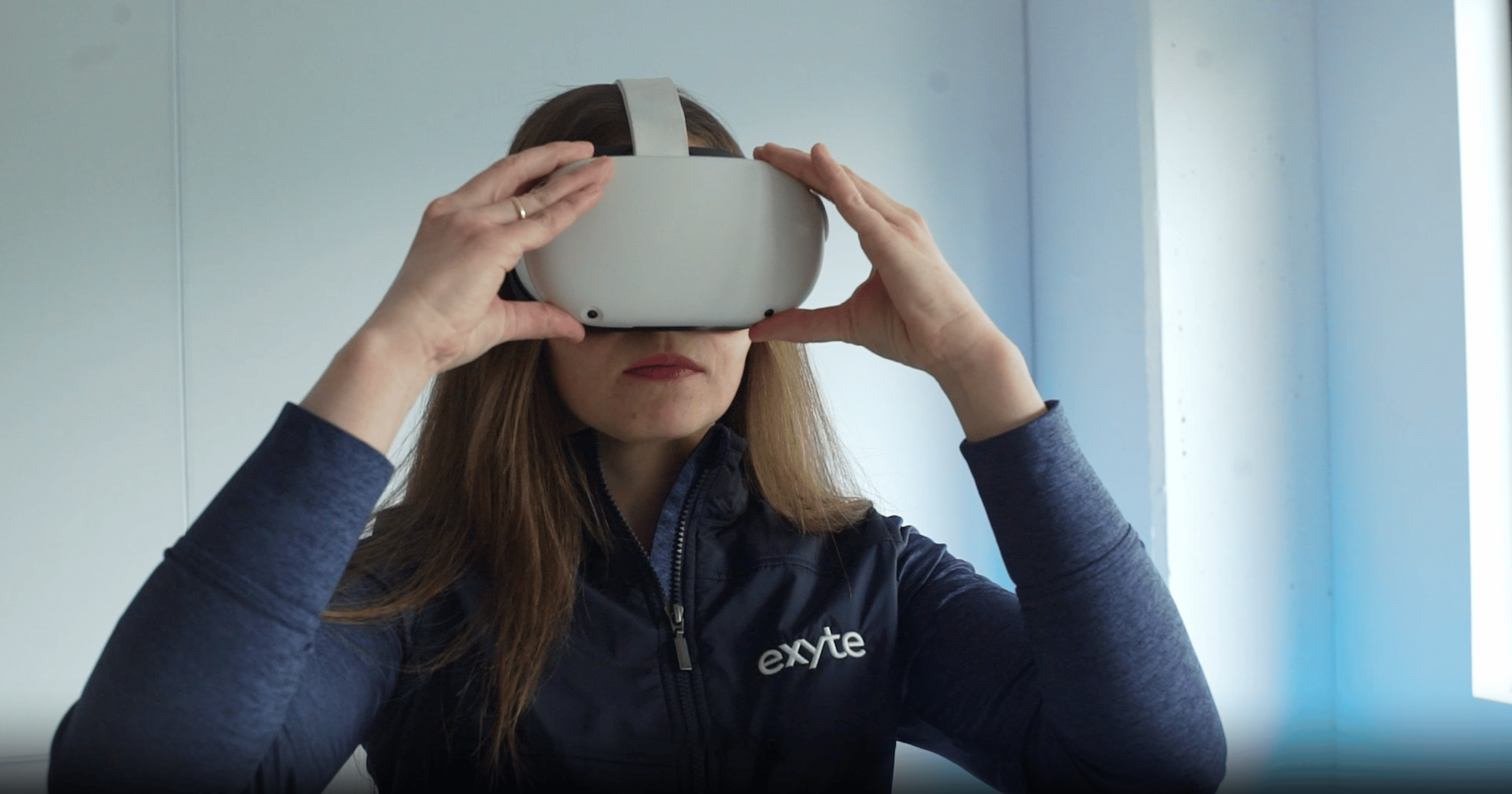 A woman wearing an Exyte-branded jacket is using a virtual reality headset, fully immersed in a digital environment.