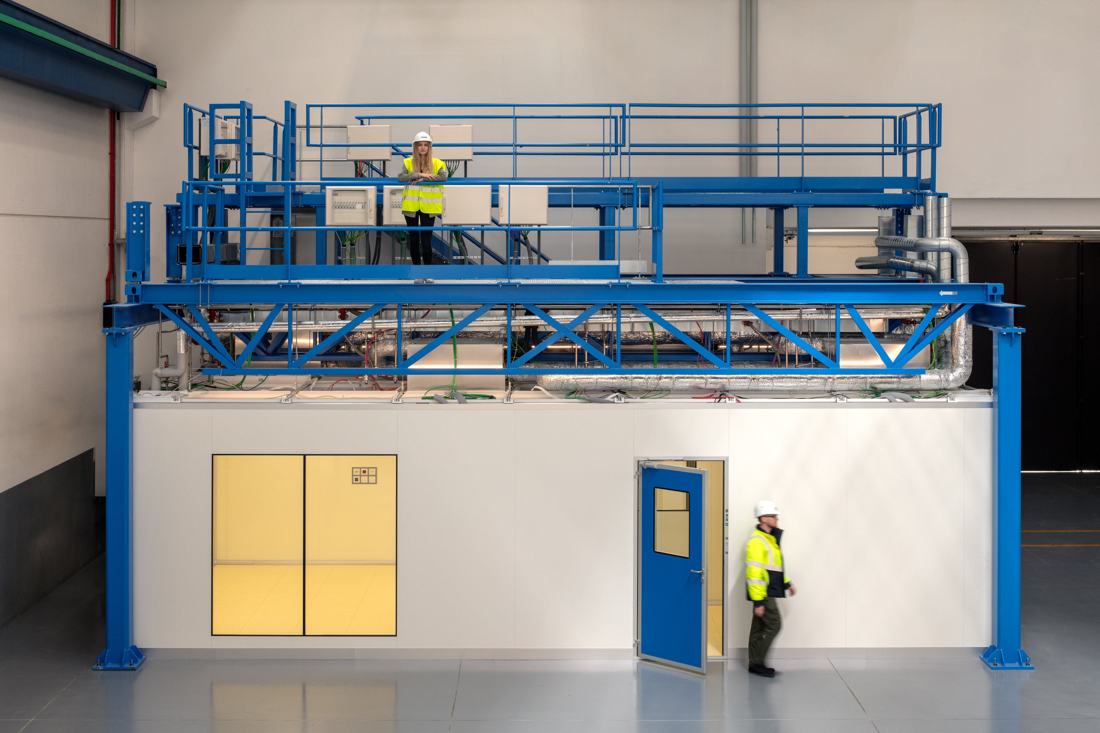 The image shows an ExyCell module, a modular cleanroom system. Two workers in safety gear are visible—one standing on an upper platform working with equipment, and another walking near the entrance door. The structure is a compact, industrial setup with blue railings and equipment on top.