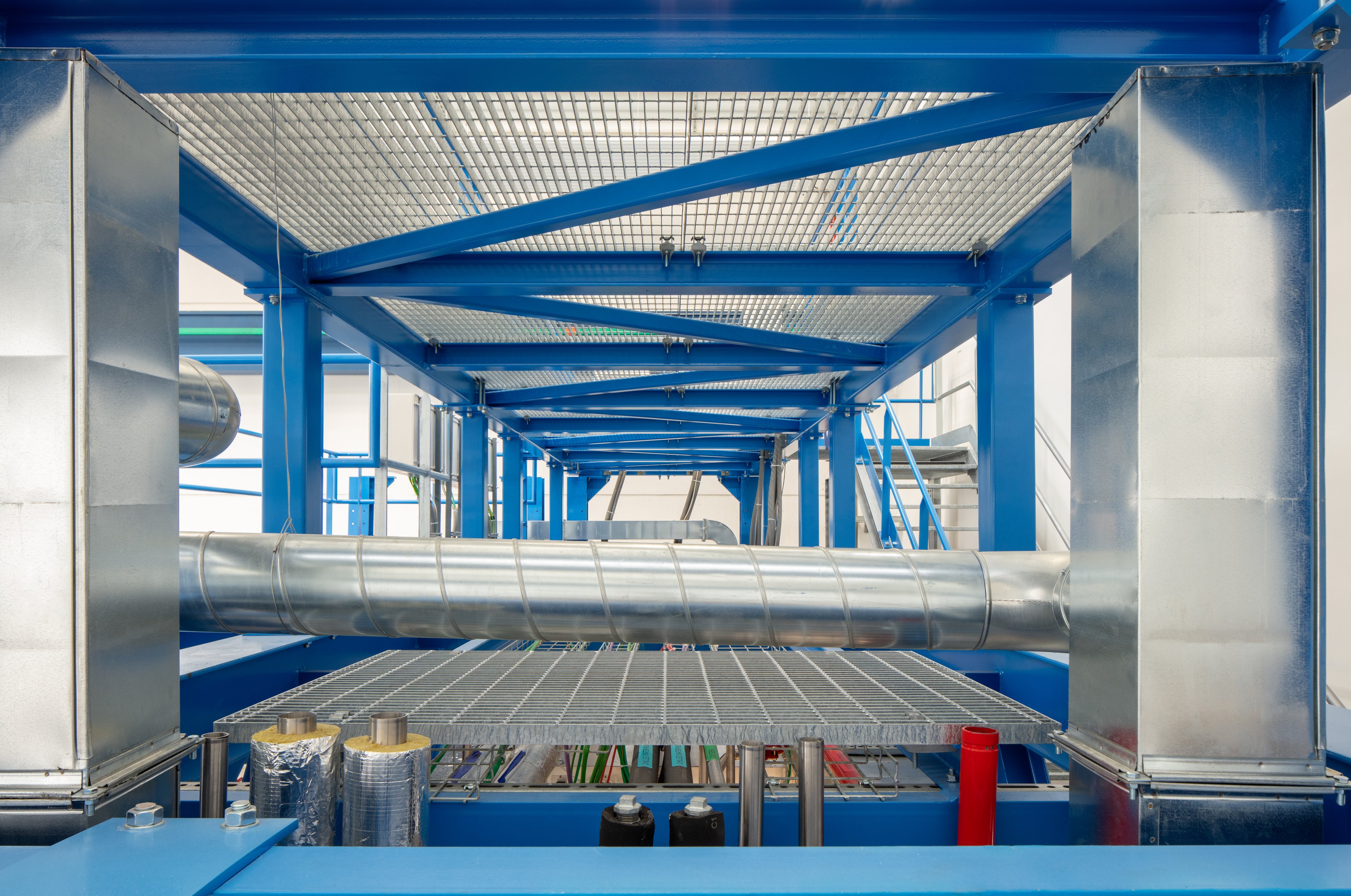 The image shows a close-up view of the ExyCell structure, highlighting a blue steel frame with metal ducting and piping running through it. The equipment setup appears to be part of a modular cleanroom or containment system, commonly used in pharmaceutical or biotech production environments. The design suggests advanced air handling and structural integrity for specialized processes.