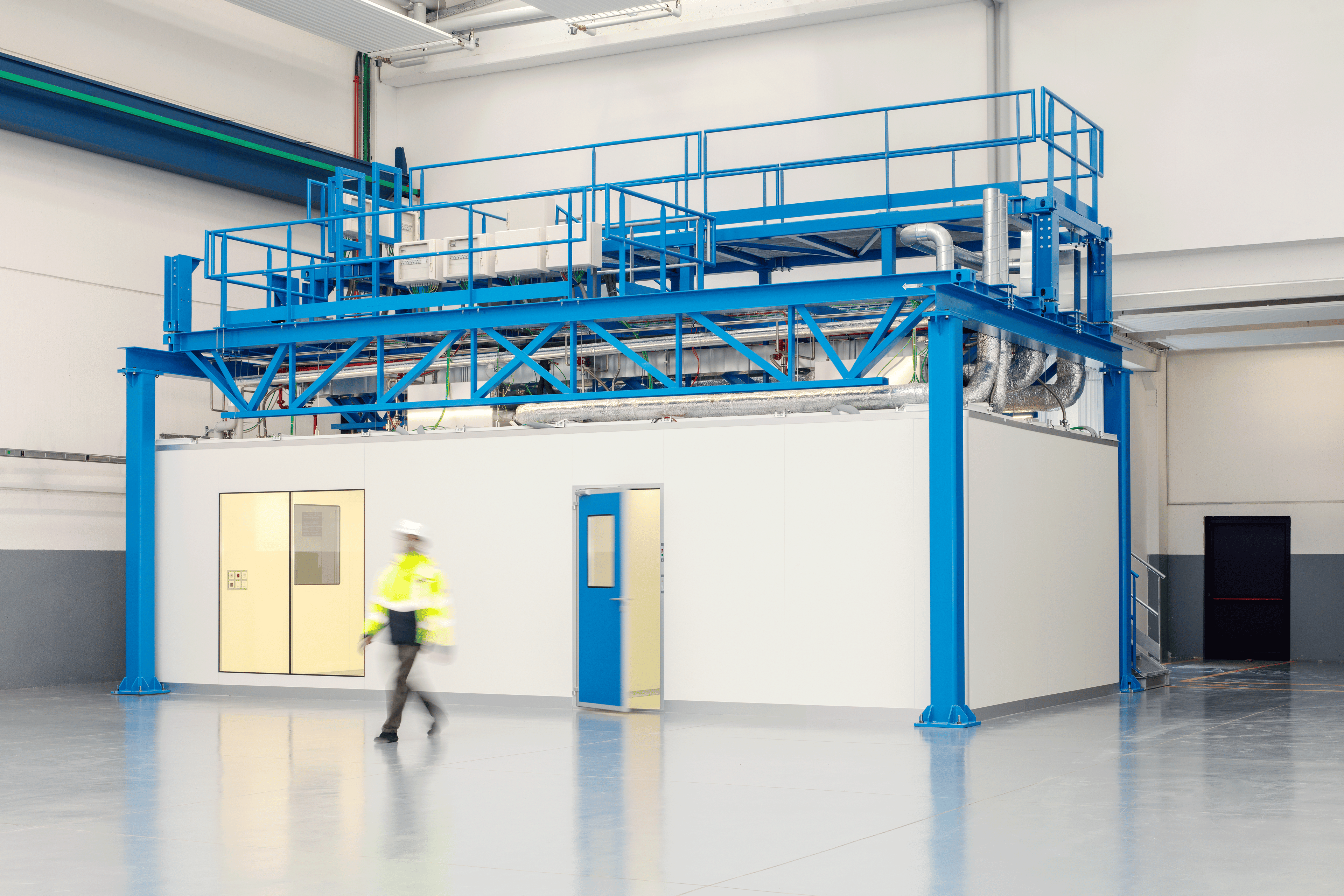 A technician in a yellow safety jacket walks past an ExyCell modular cleanroom structure, featuring a blue framework and piping systems above the enclosed unit in a large industrial facility.