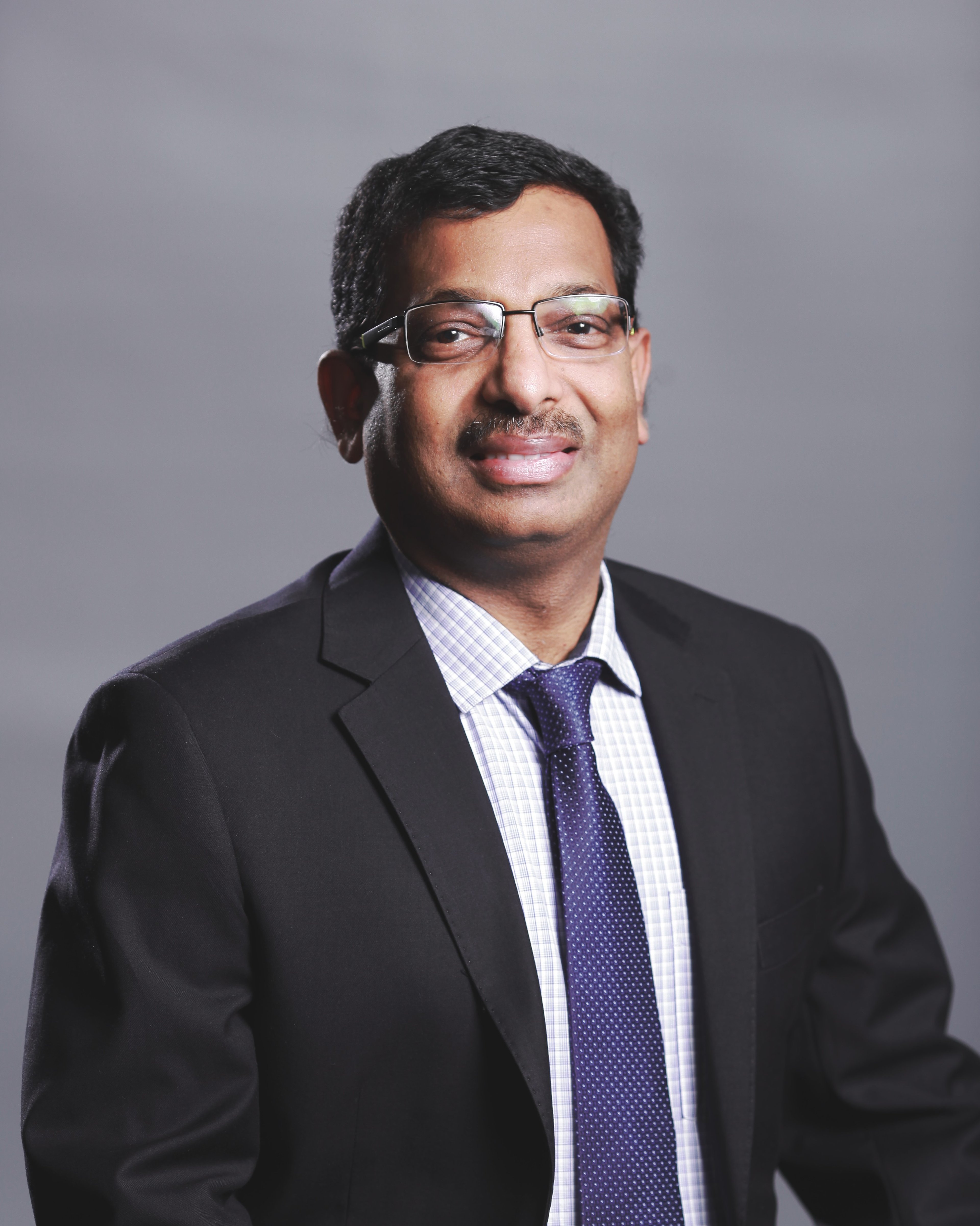 A portrait of Maheswaran Nair, wearing glasses, a dark suit, checkered shirt, and a navy tie, with a neutral grey background.
