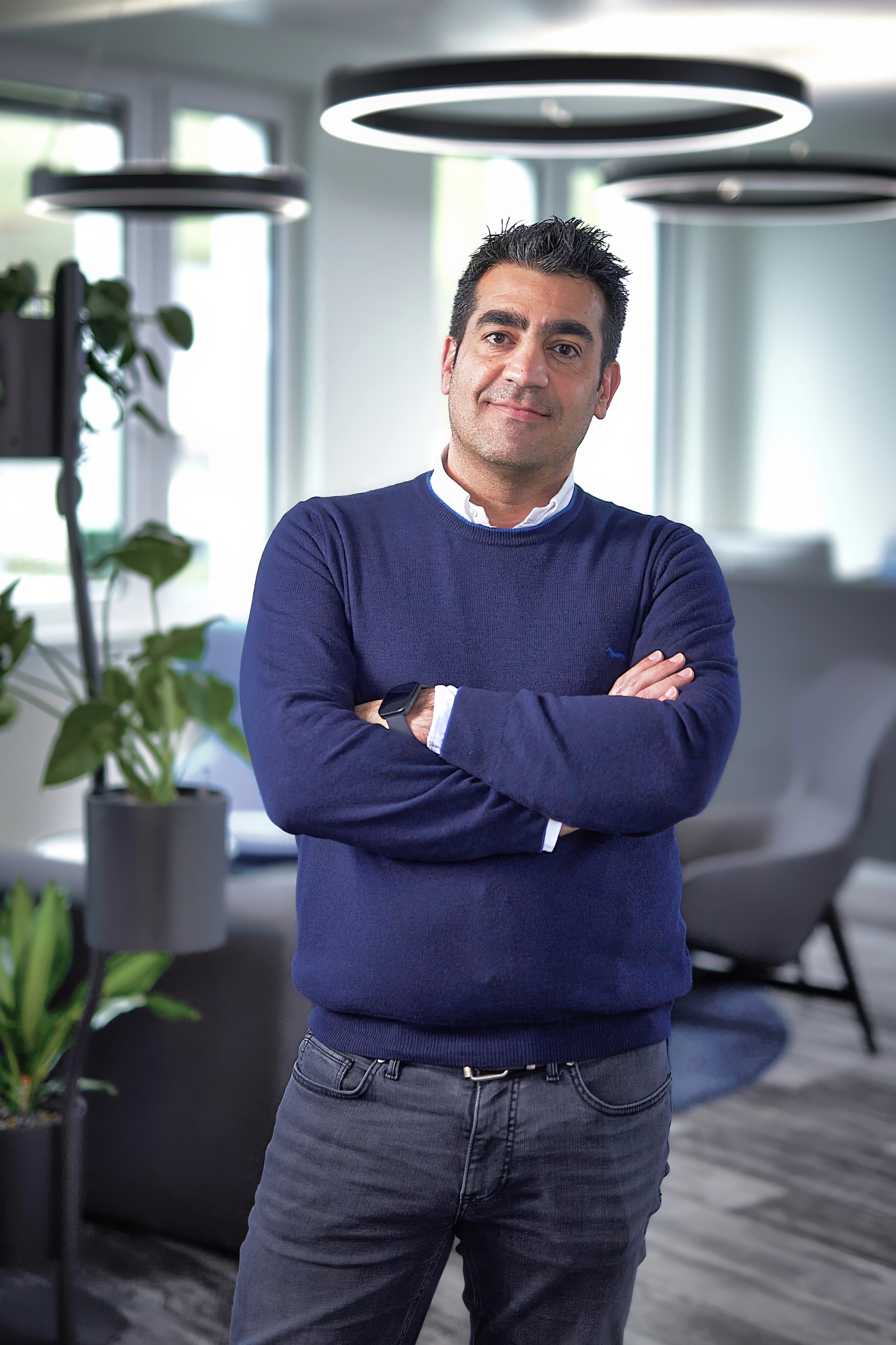  Antonello Finucci stands confidently with his arms crossed in a modern office setting. He wears a navy blue sweater and dark jeans, exuding a relaxed yet professional demeanor.