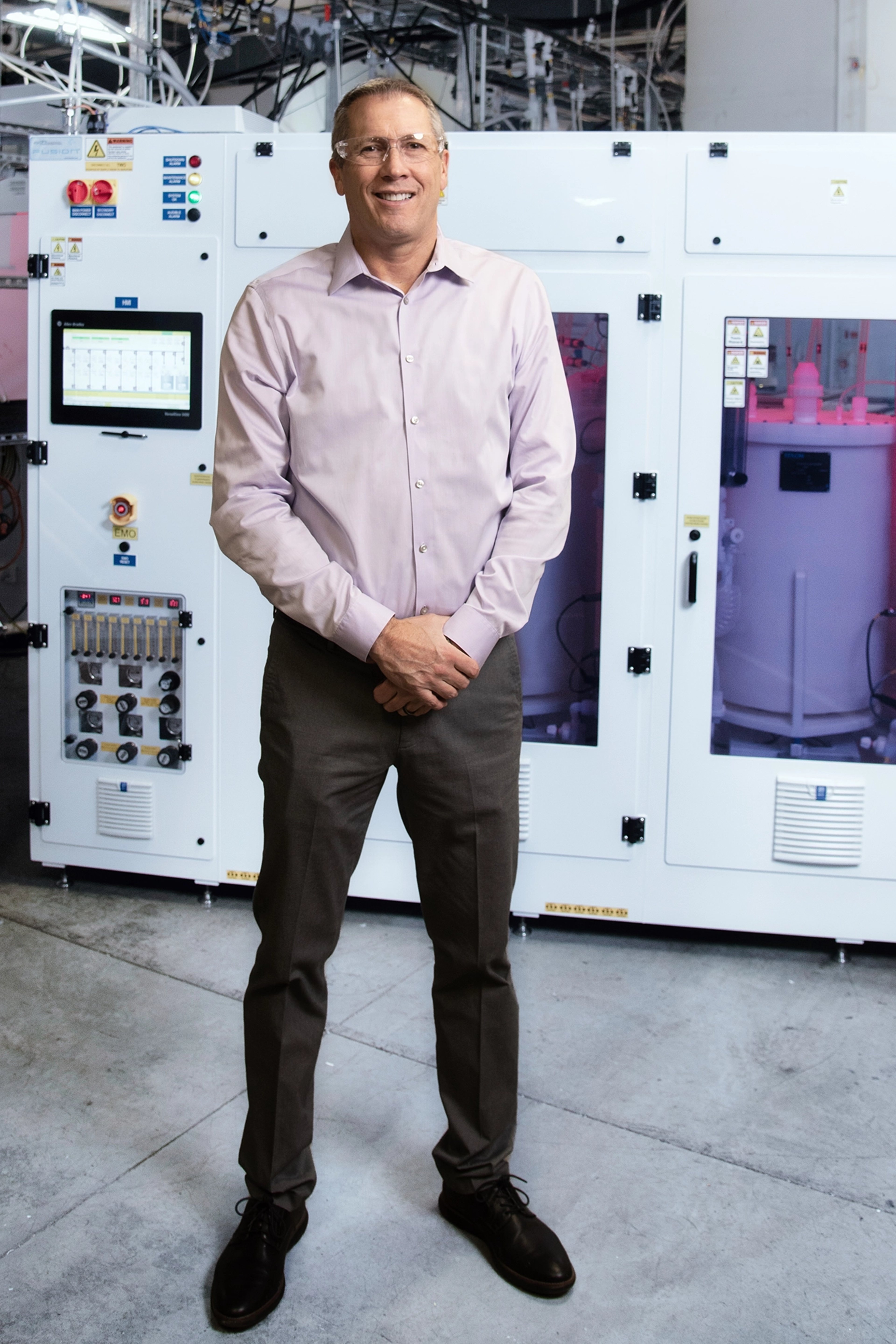 Darren Willey stands in front of advanced industrial equipment, dressed in a light pink shirt and dark trousers, smiling confidently.