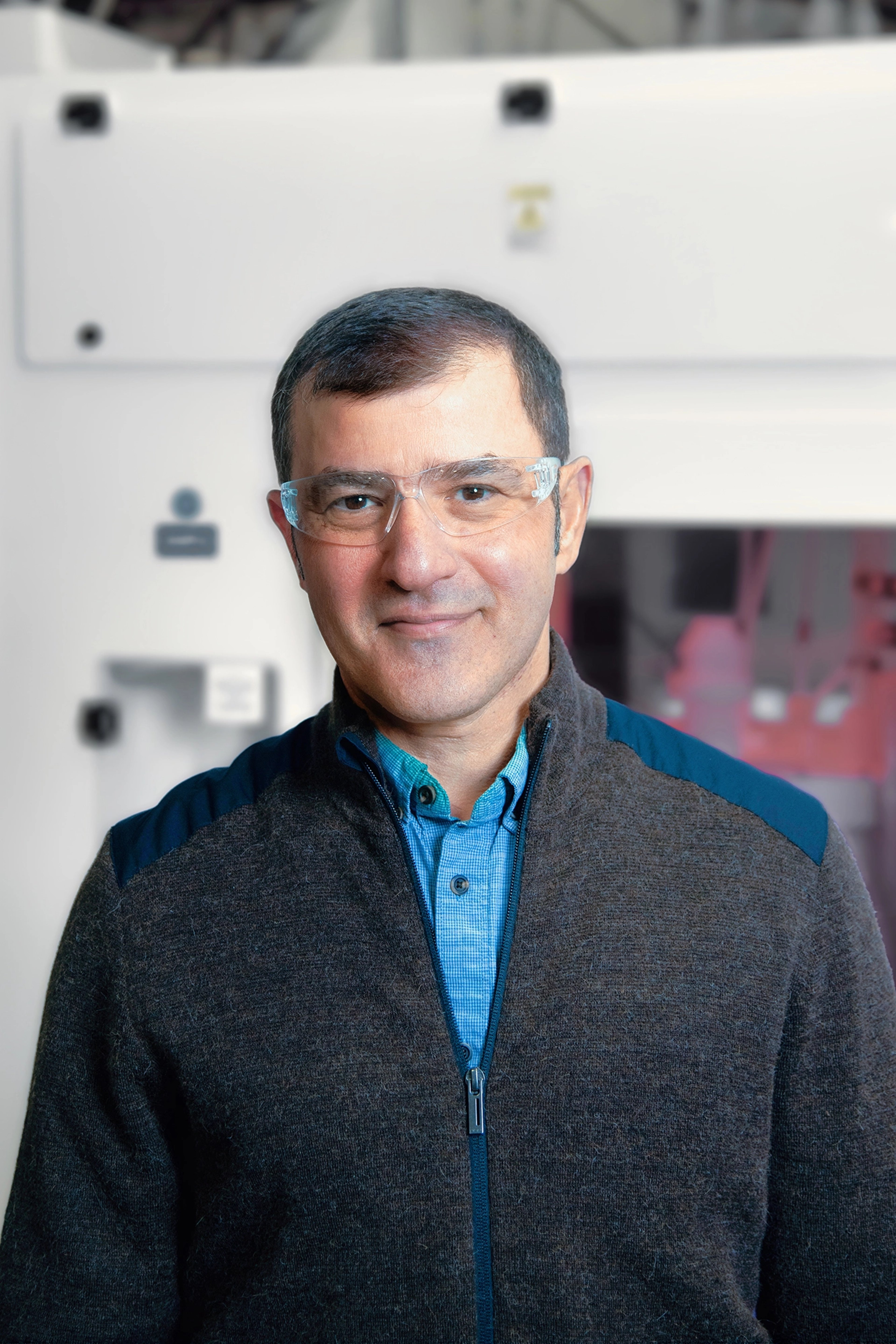 Dawood Abugharbieh is wearing safety glasses, a blue shirt, and a dark jacket, standing in front of advanced industrial equipment with a composed expression.