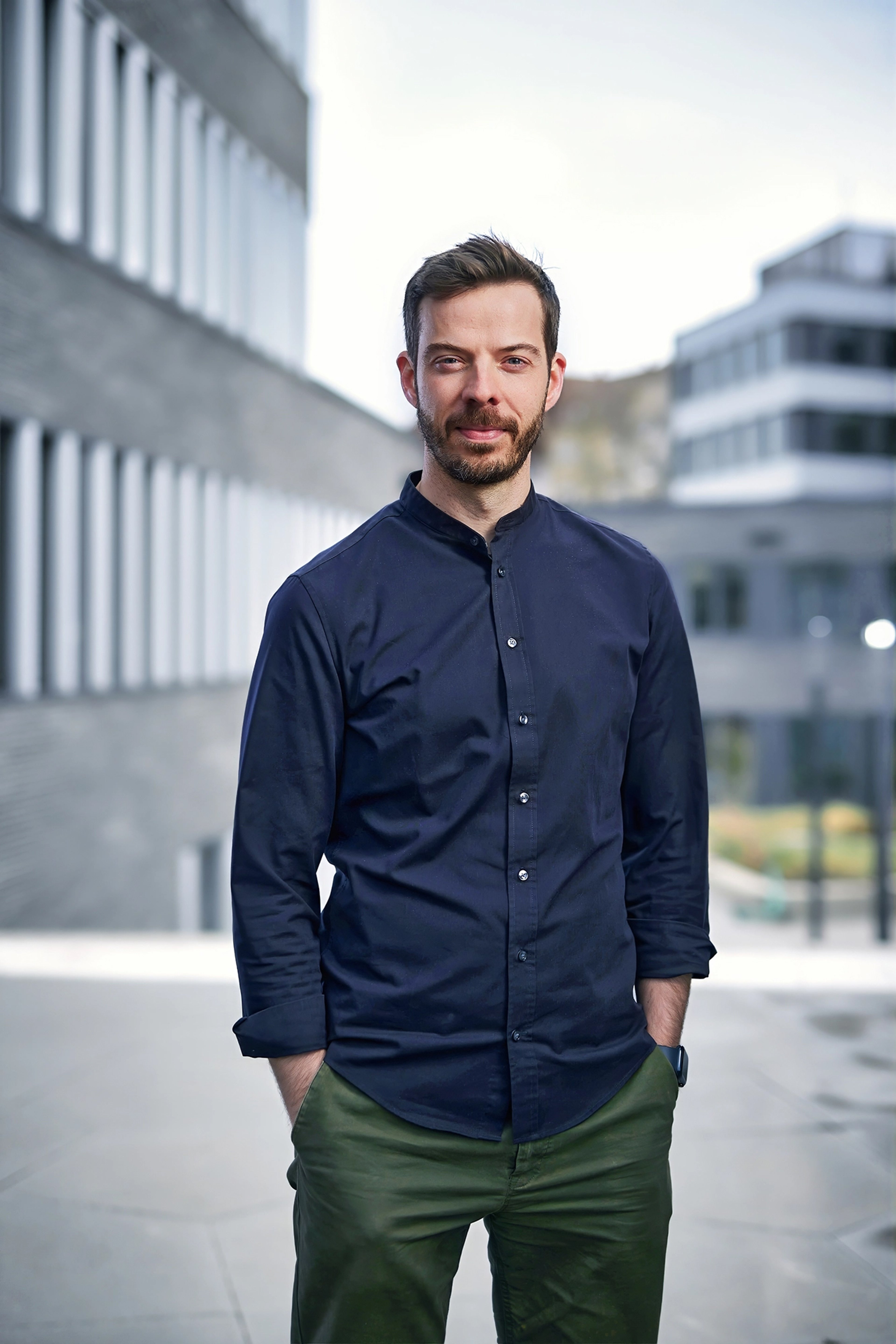 Julian Kuczaty is standing outside, wearing a dark blue buttoned shirt and olive green pants. He faces the camera with his hands in his pockets, set against a modern office building background.