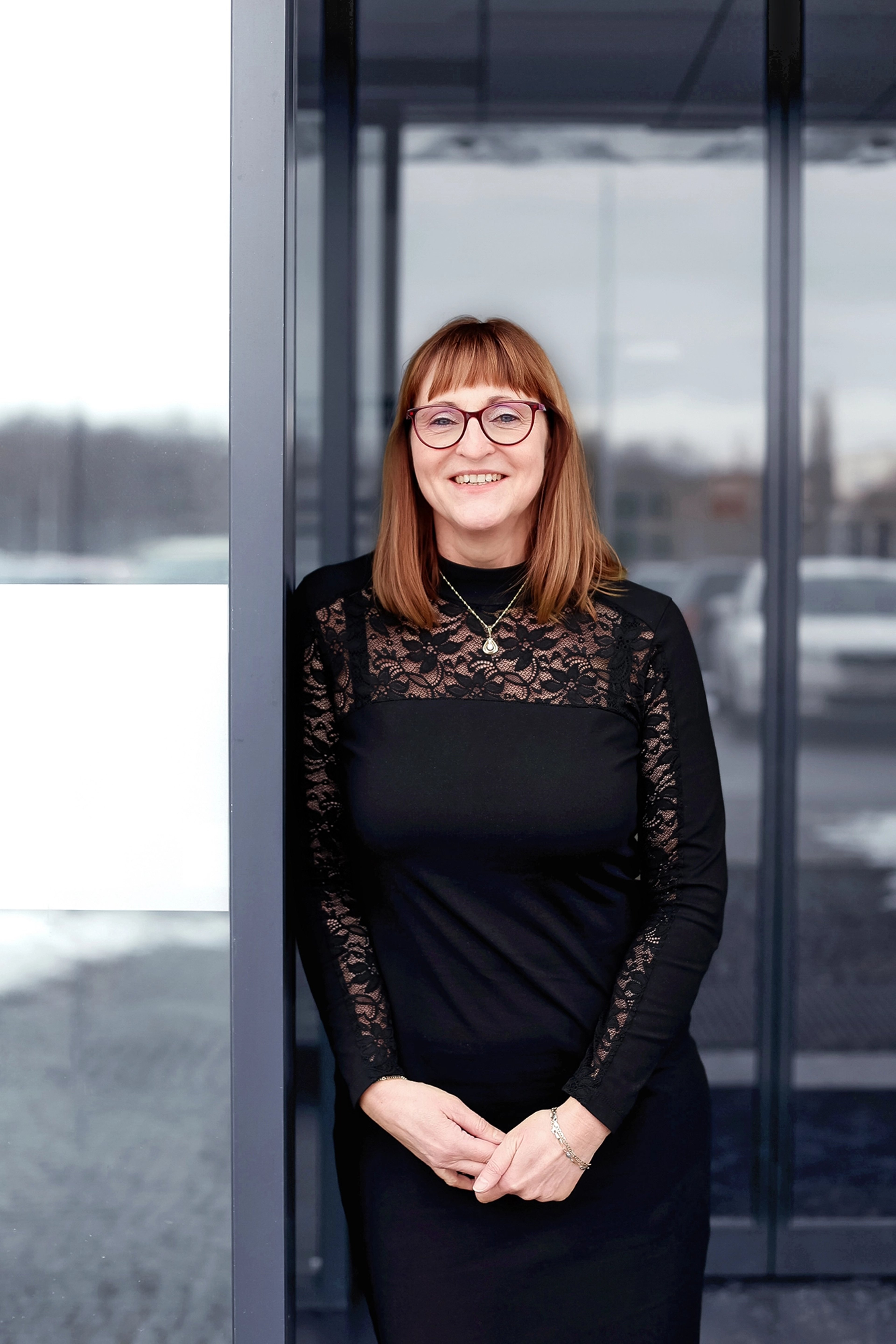 Lucie Weberova is standing near a glass entrance, wearing glasses and a black lace-detailed dress. She smiles confidently with her hands gently clasped in front of her.