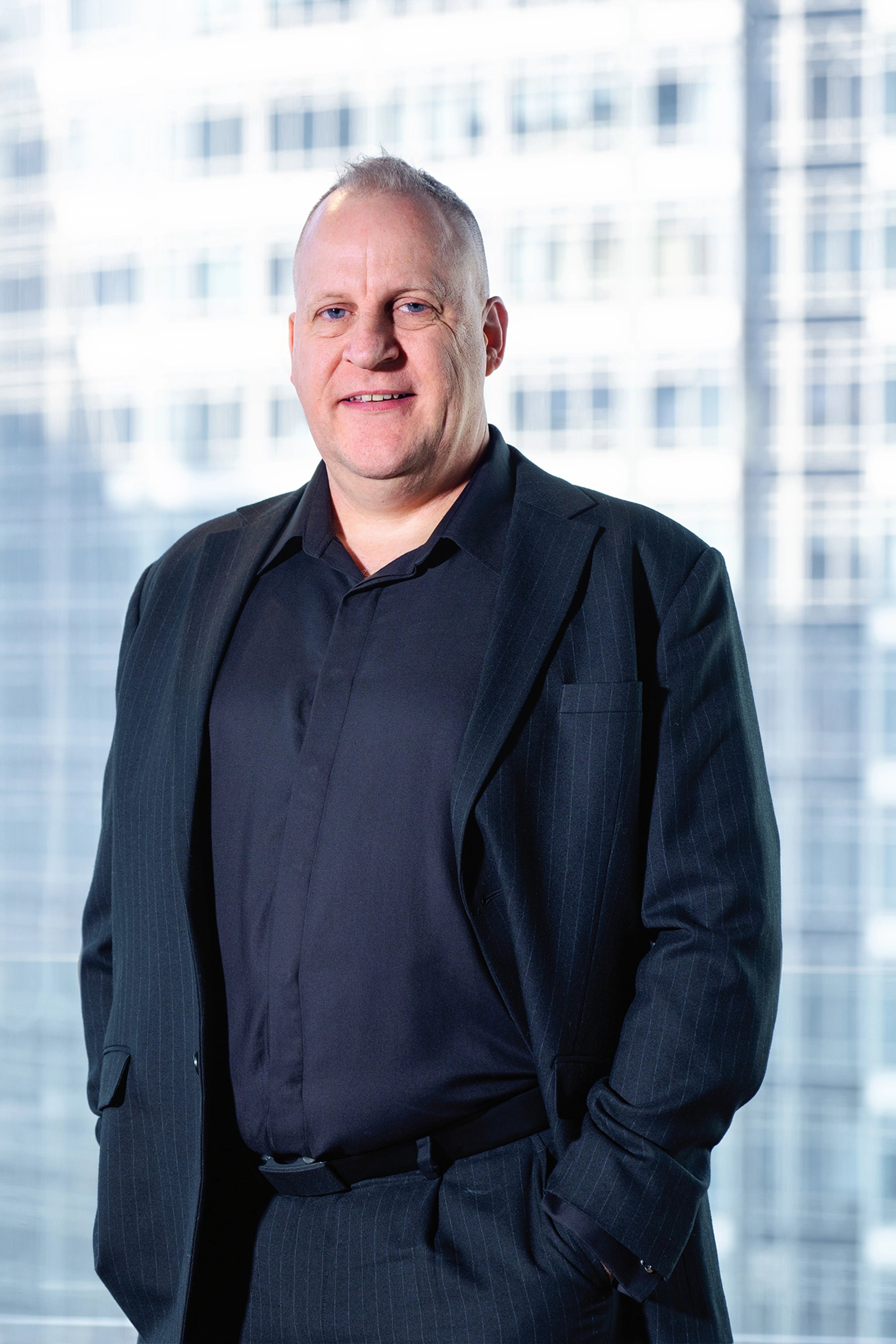 Mark Stephens stands in front of a bright backdrop of office windows, wearing a dark suit and shirt, with his hands casually in his pockets. He smiles slightly, exuding a professional yet approachable demeanor.
