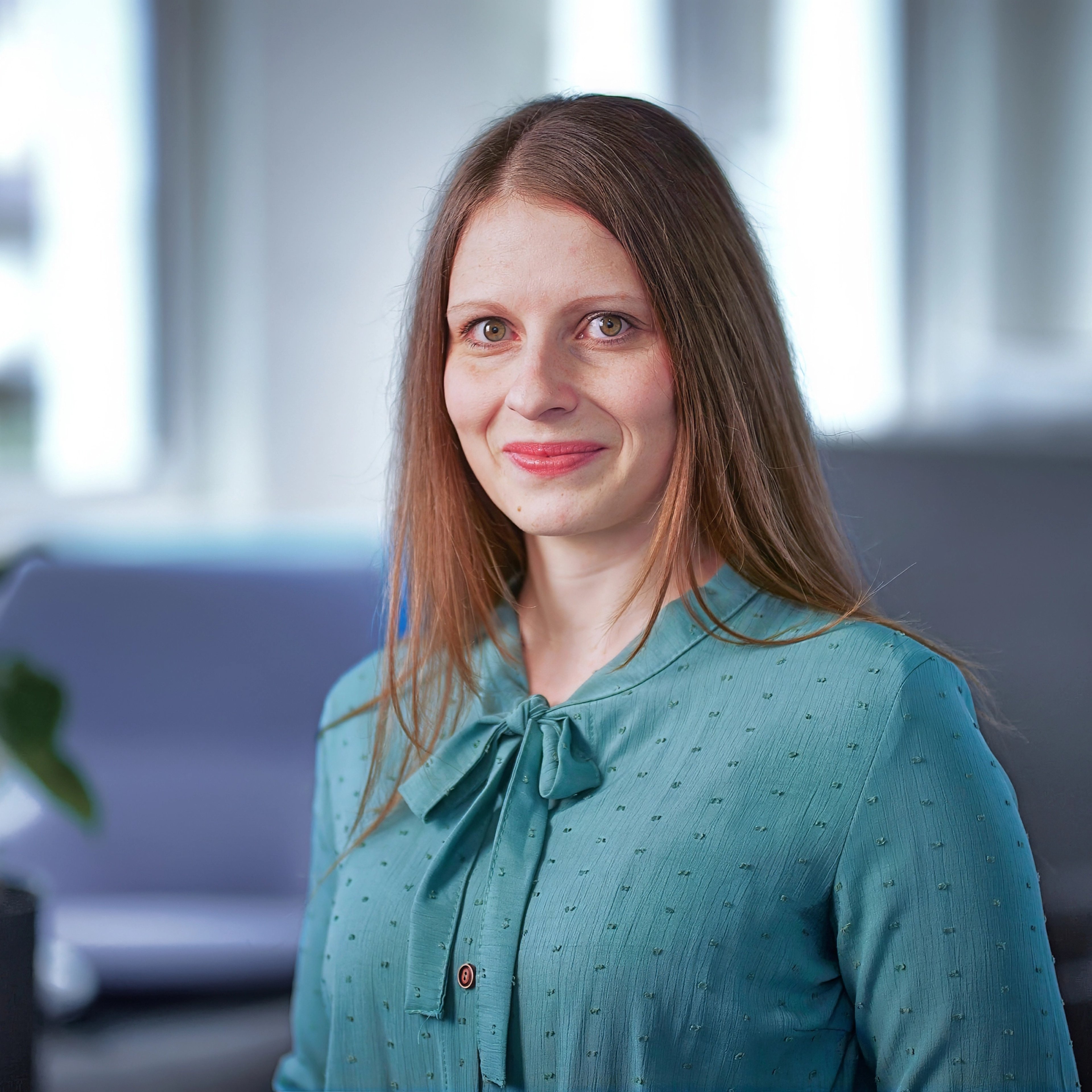 Olga Fischbein smiles softly while standing indoors. She wears a teal blouse with a bow at the neck, her long brown hair gently resting on her shoulders. The background is softly lit, creating a calm and welcoming atmosphere.