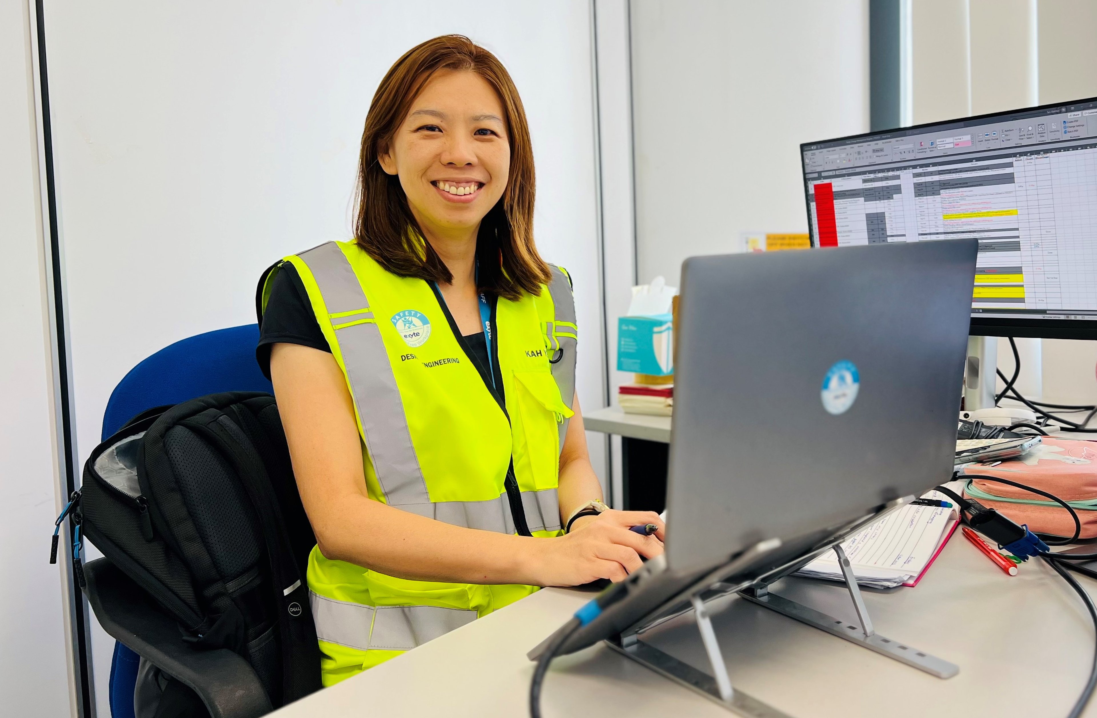 Tan Kah Yen, wearing a high-visibility Exyte vest, is sitting at a desk working on a laptop. She is smiling, with an open spreadsheet displayed on a monitor beside her.