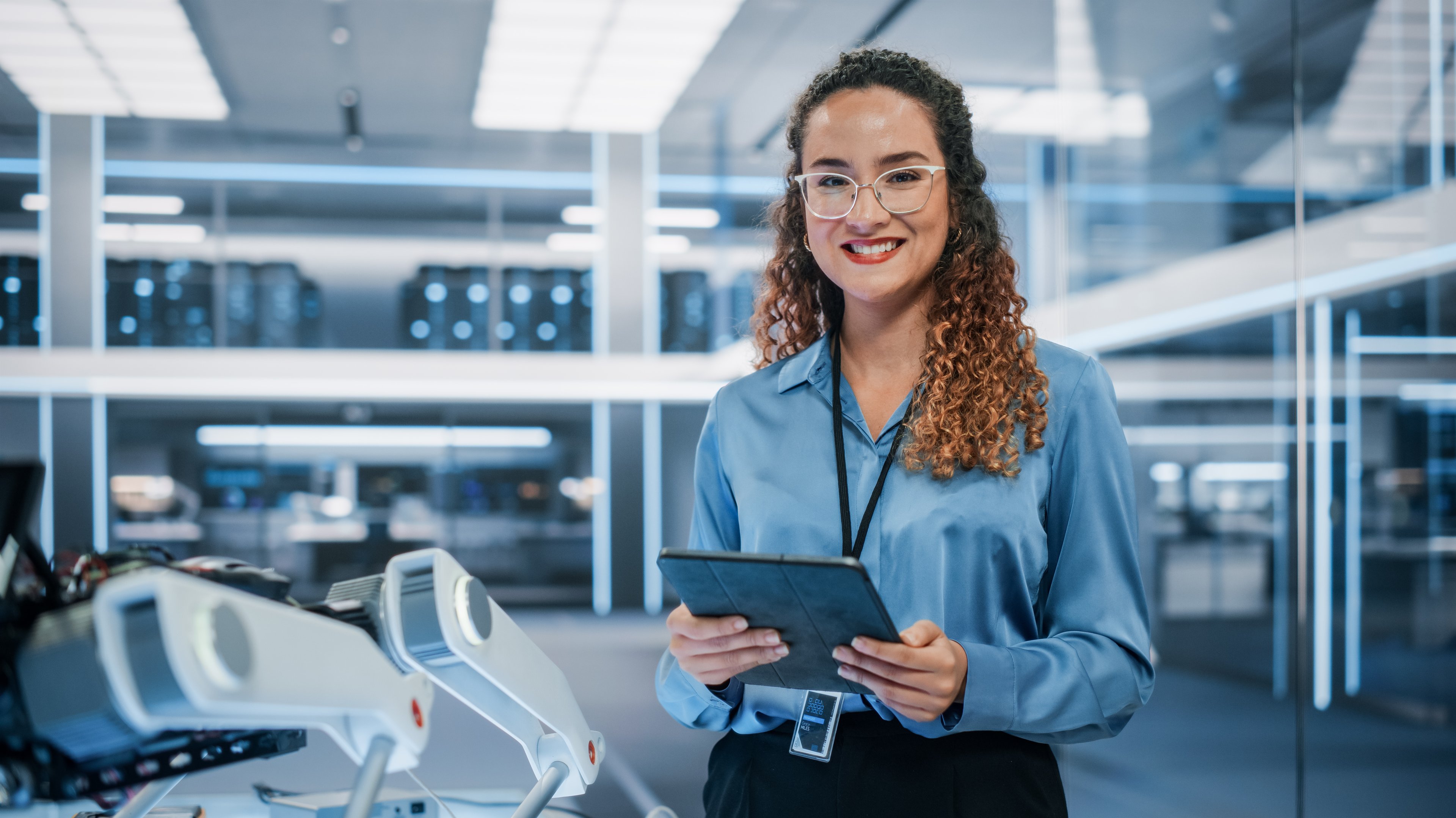 A woman standing in a modern technology lab, smiling while holding a tablet. She is wearing glasses and a blue blouse, with advanced robotic equipment visible in the background.