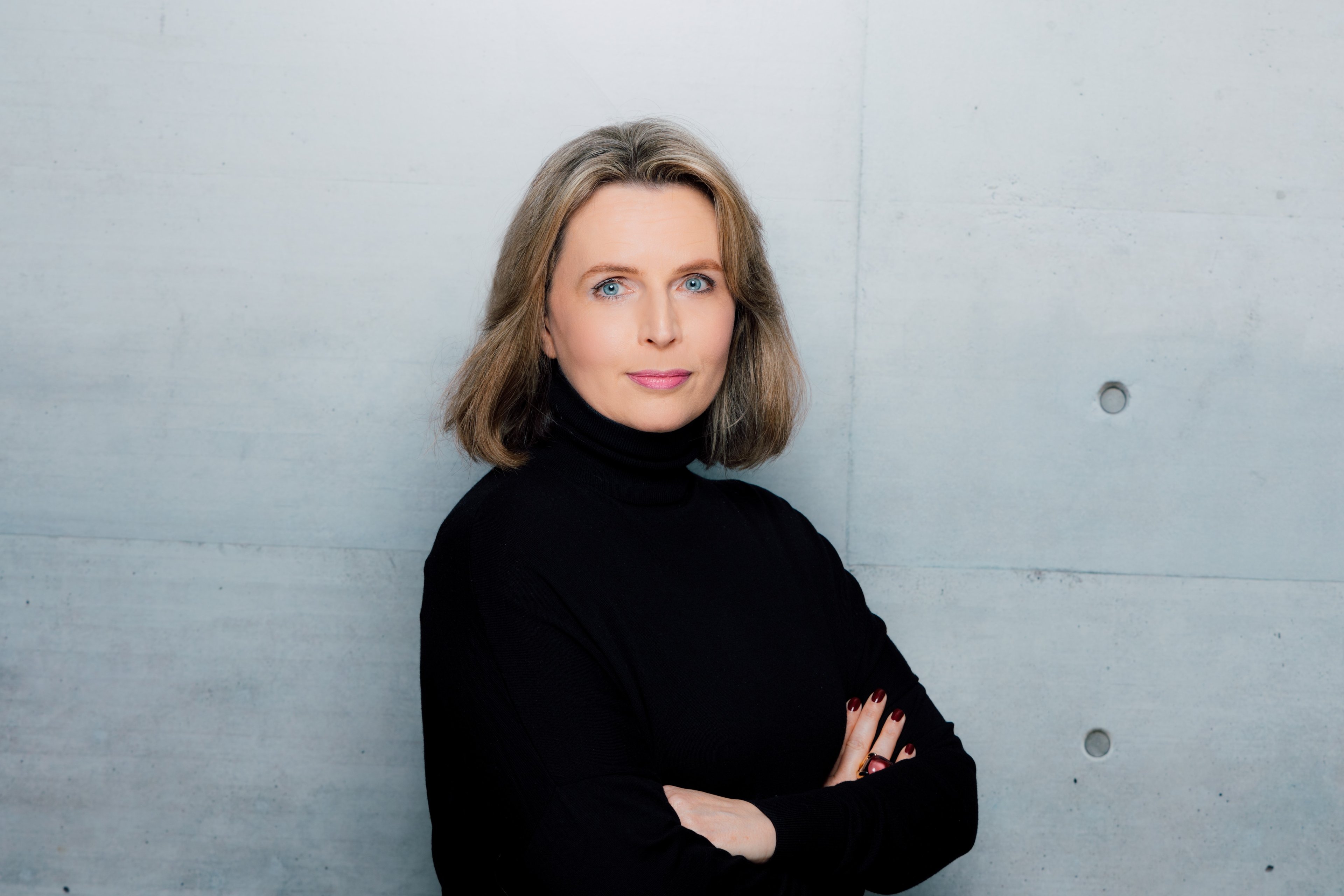 A professional portrait of Alexandra Kuebler, standing confidently with arms crossed, wearing a black turtleneck, in front of a modern, minimalist concrete wall.