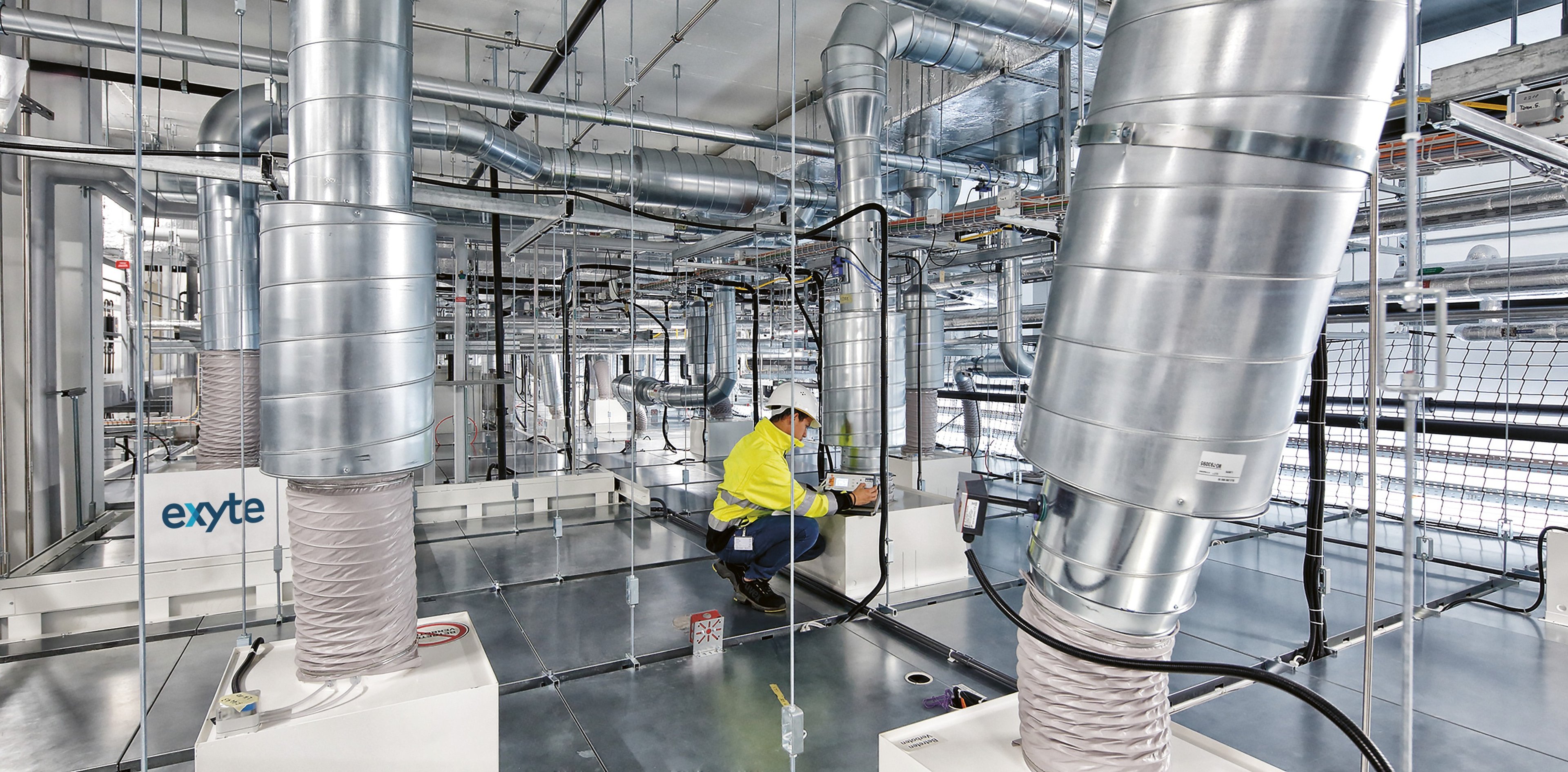 A technician working on advanced HVAC systems in a cleanroom environment with the Exyte logo visible, surrounded by complex ductwork and equipment, ensuring air quality and ventilation for precision manufacturing.