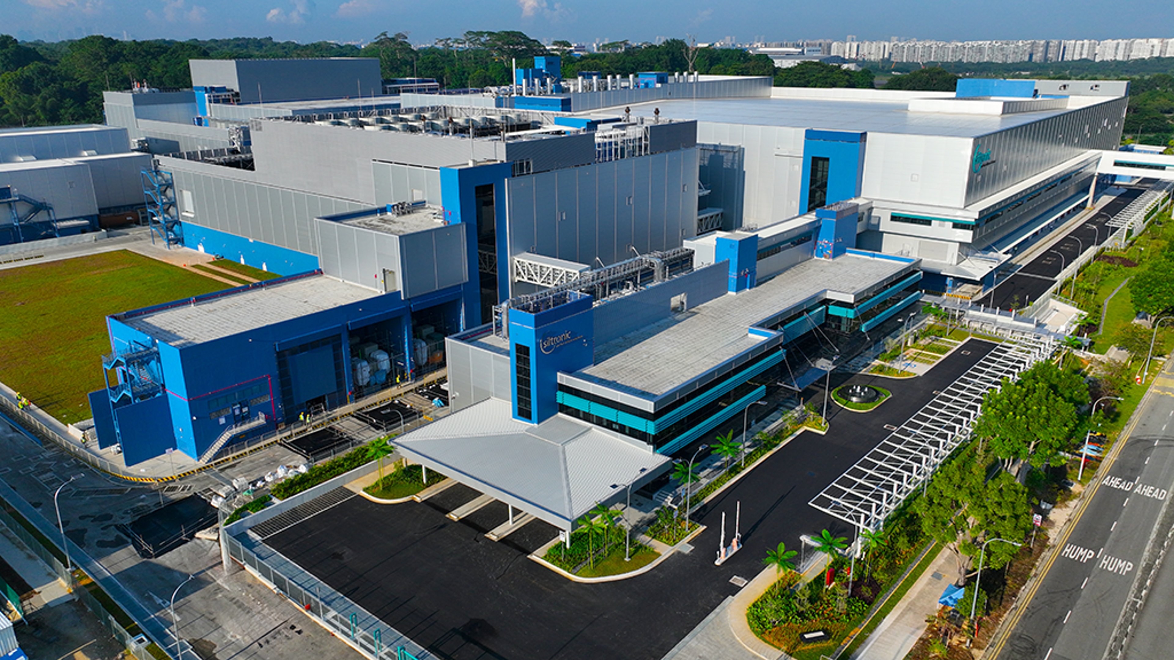 An aerial view of the Siltronic semiconductor fab project in Singapore, showcasing large industrial buildings with blue accents and surrounding infrastructure, including roads and greenery.