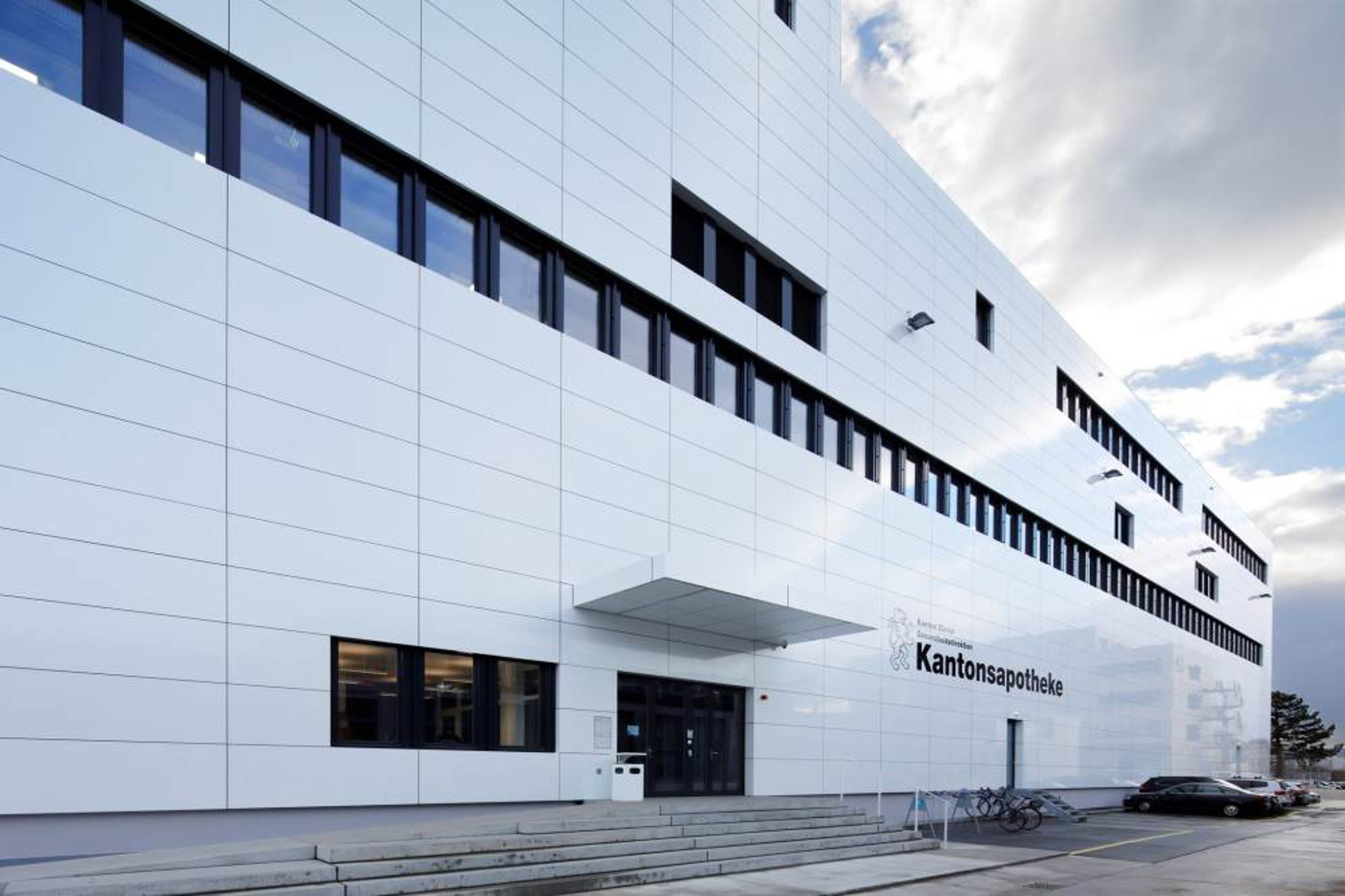 An exterior view of the Kantonsapotheke Zurich, a producing pharmacy for hospitals located in Schlieren, featuring a sleek modern white facade with black-framed windows.