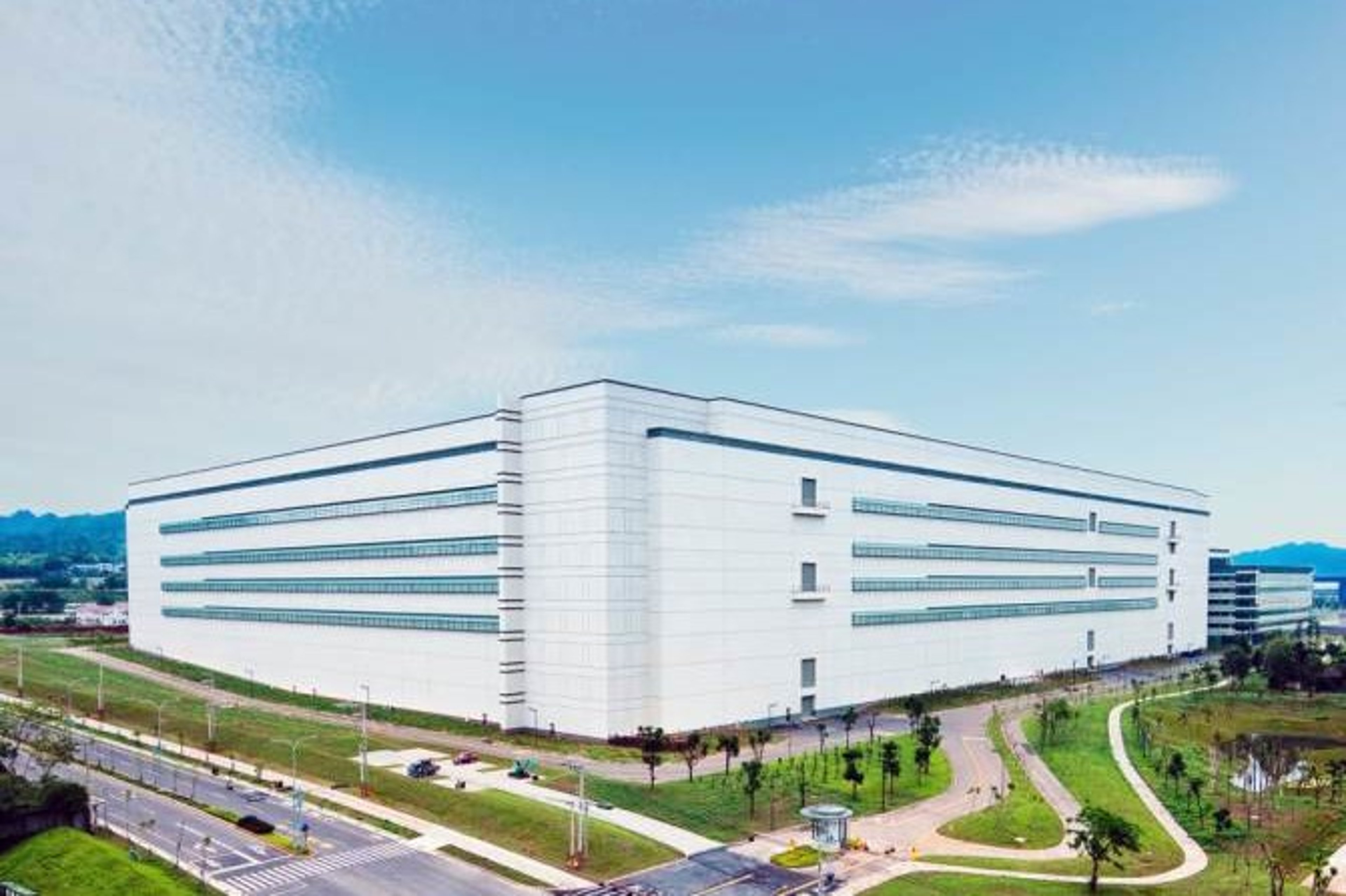 An exterior view of the Gen. 8.5 FPD production facility expansion in Houli, featuring a large, modern white building with multiple levels, surrounded by green landscaping and roadways.