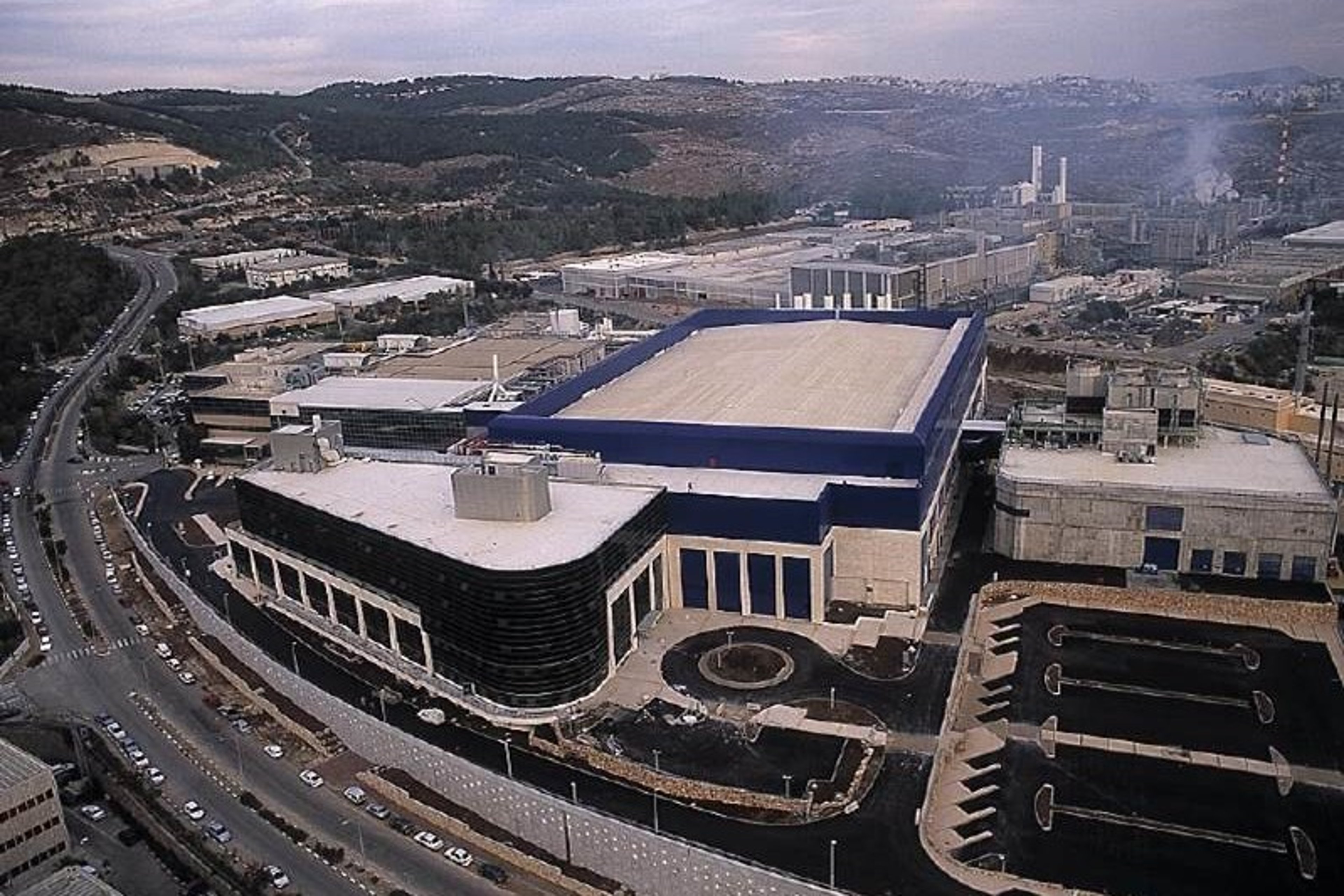 An aerial view of a semiconductor fab in Migdal Ha’Eemek, featuring large industrial buildings surrounded by roads and set against a backdrop of hills and greenery.