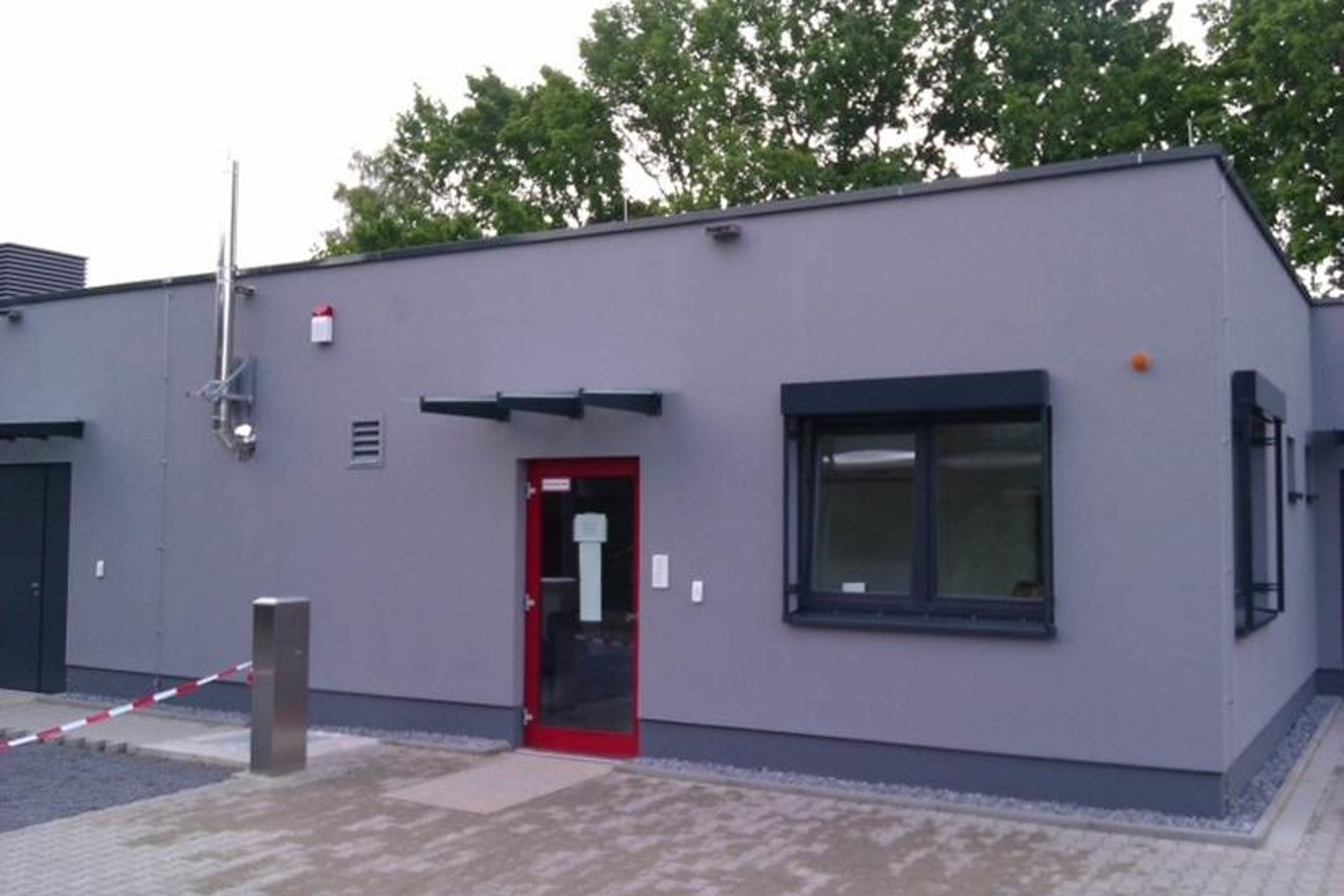 An exterior view of a pharma lab and production facility in Gütersloh, featuring a modern grey building with a red entrance door and surrounding greenery.