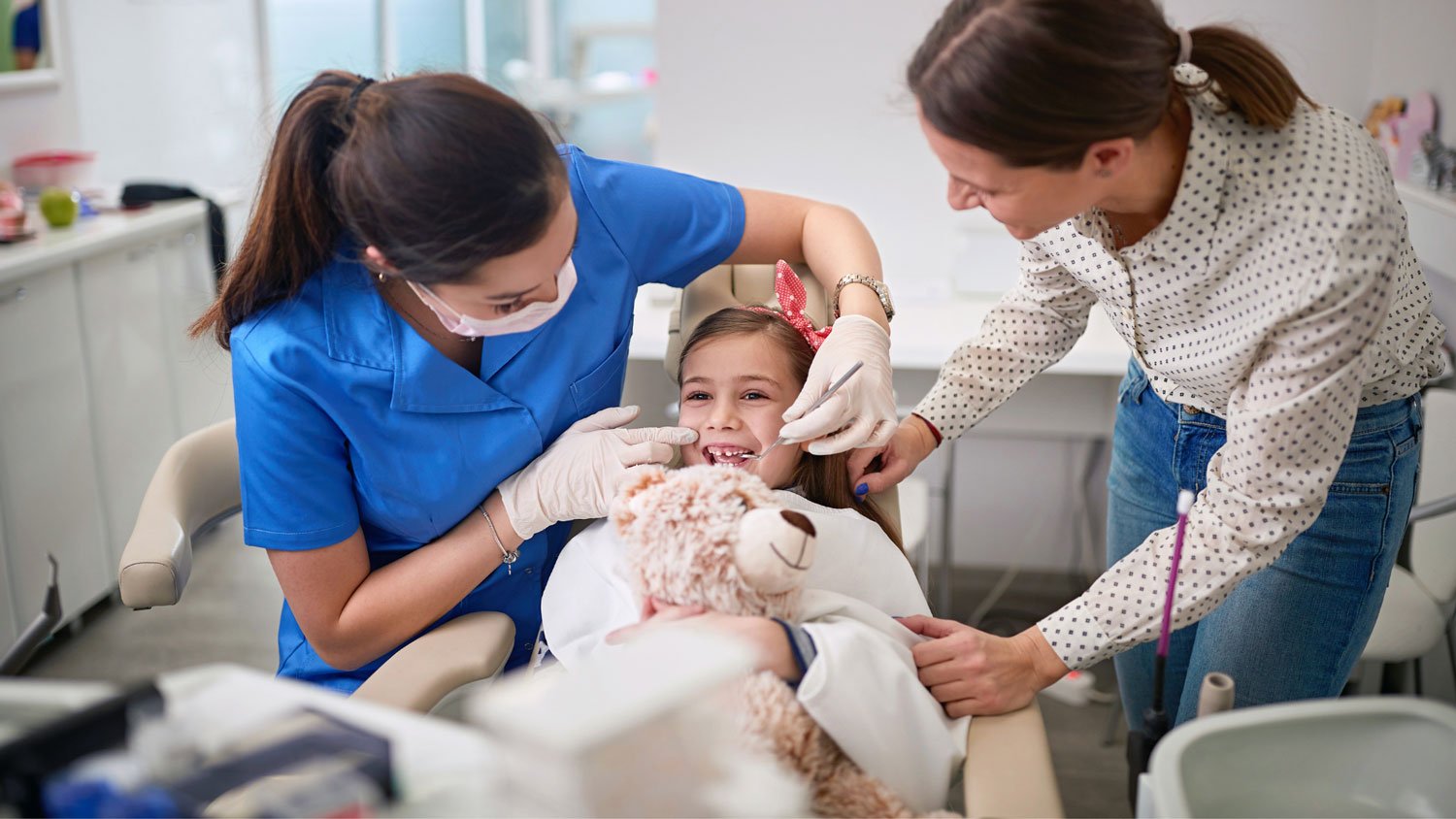 A young girl with a teddy bear being attended by a dentist and her mother in a dental clinic 