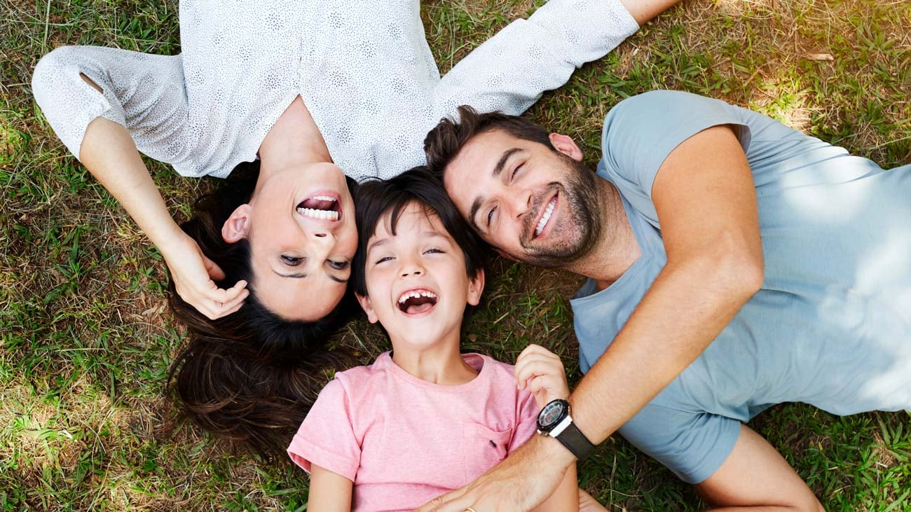 A family of three, a mother, father, and young son, lying head-to-head on the grass, laughing joyfully under a clear sky