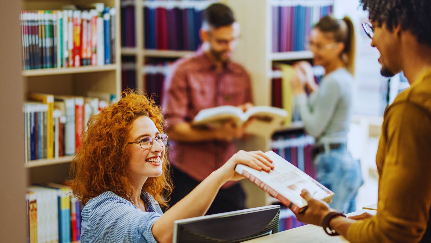 A student handing a book back to a librarian with two students reading a book together in the background