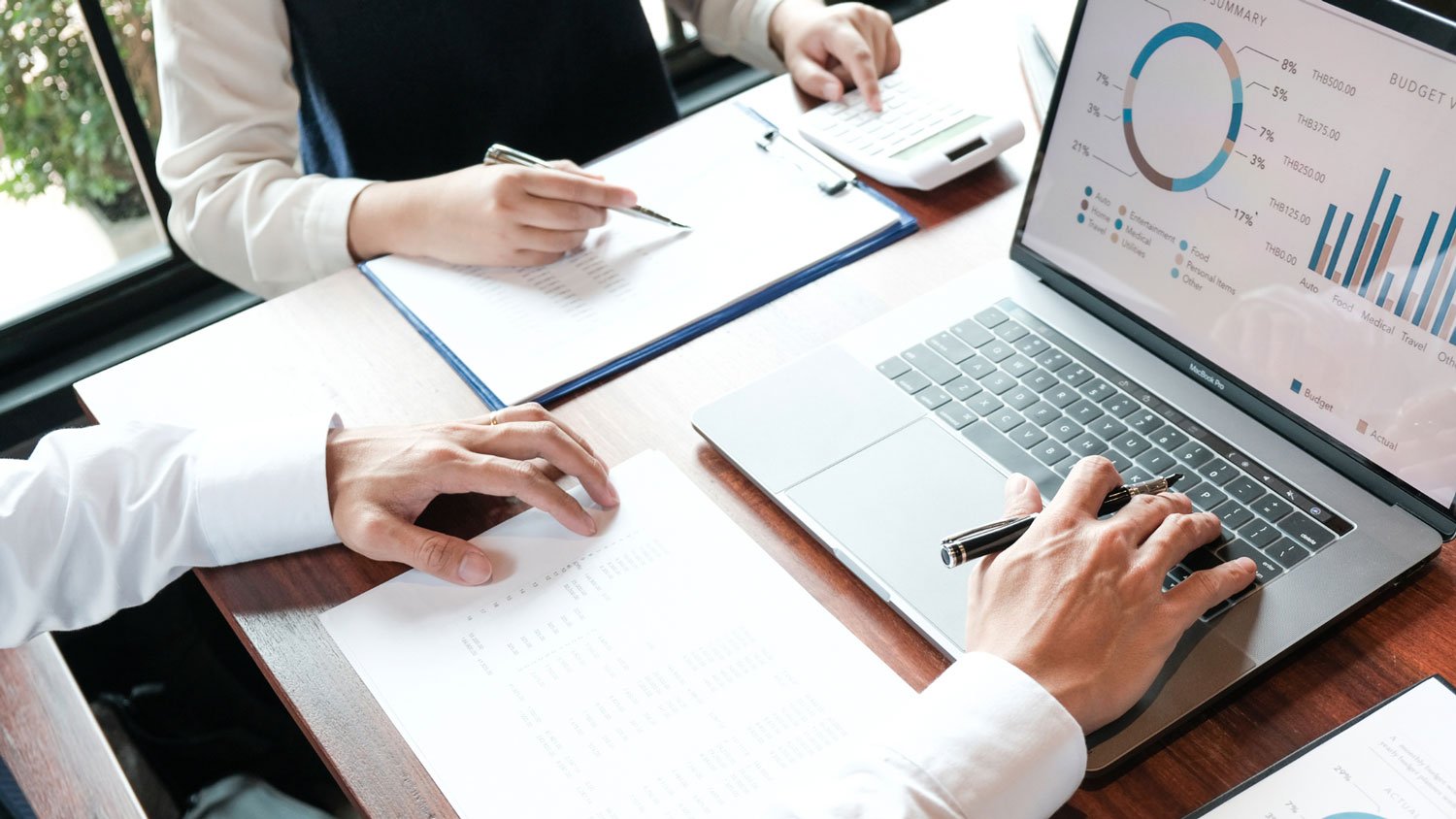 Two professionals reviewing financial documents on a laptop at a sunlit desk.