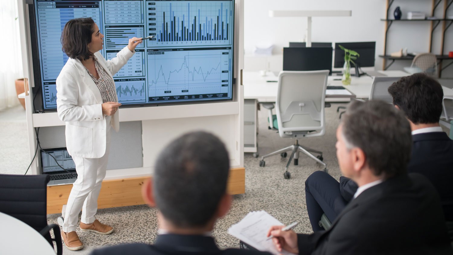 A woman presenting financial data on large screens to an audience.