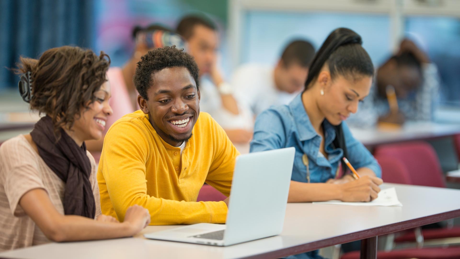 A classroom scene with diverse students smiling and interacting around a laptop, emphasizing collaboration and enjoyment in learning