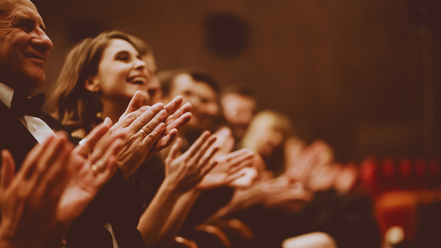Audience members in formal attire clapping enthusiastically in a dimly lit theatre