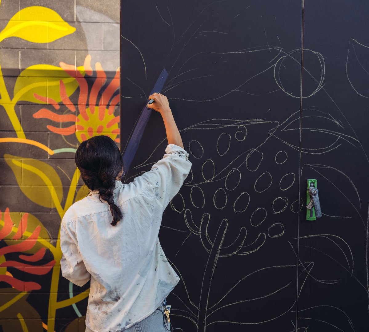A woman artist sketching floral patterns on a large black canvas outdoors, with colorful designs in the background