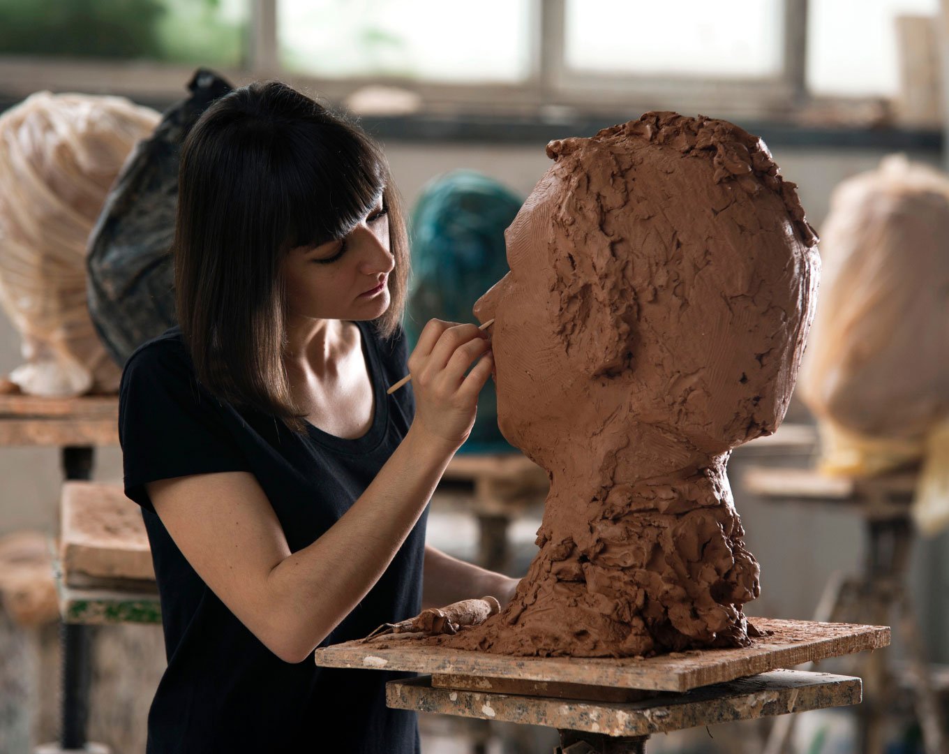 A female sculptor carefully modeling a clay bust in a bright, sunlit studio surrounded by various sculptures
