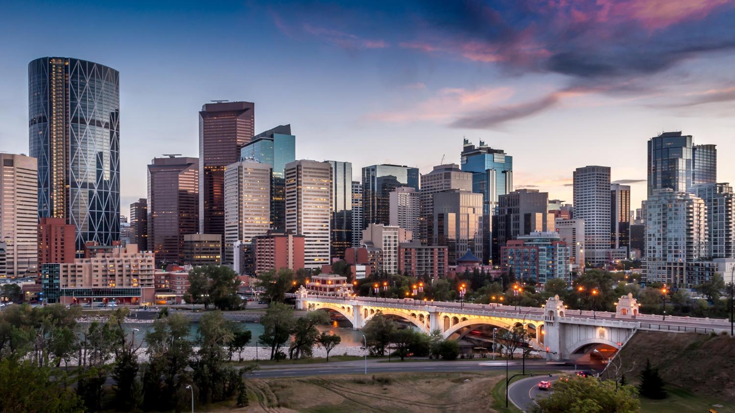 A panoramic view of the Calgary skyline at sunset, showing rivers and a bridge.