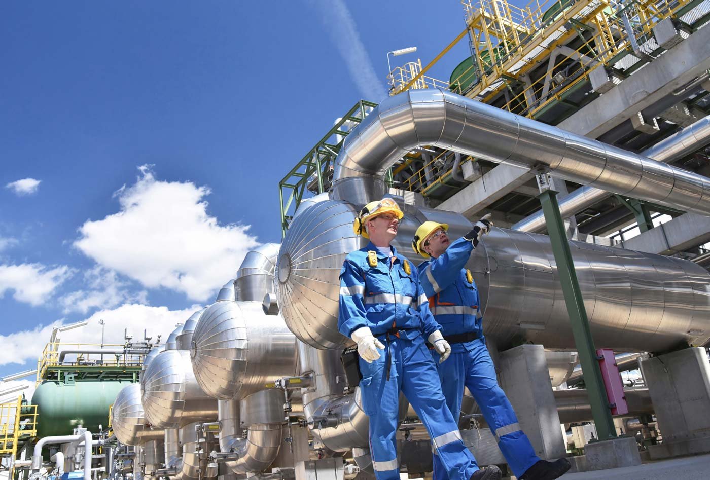Two workers in blue overalls discussing over industrial pipelines at a large plant
