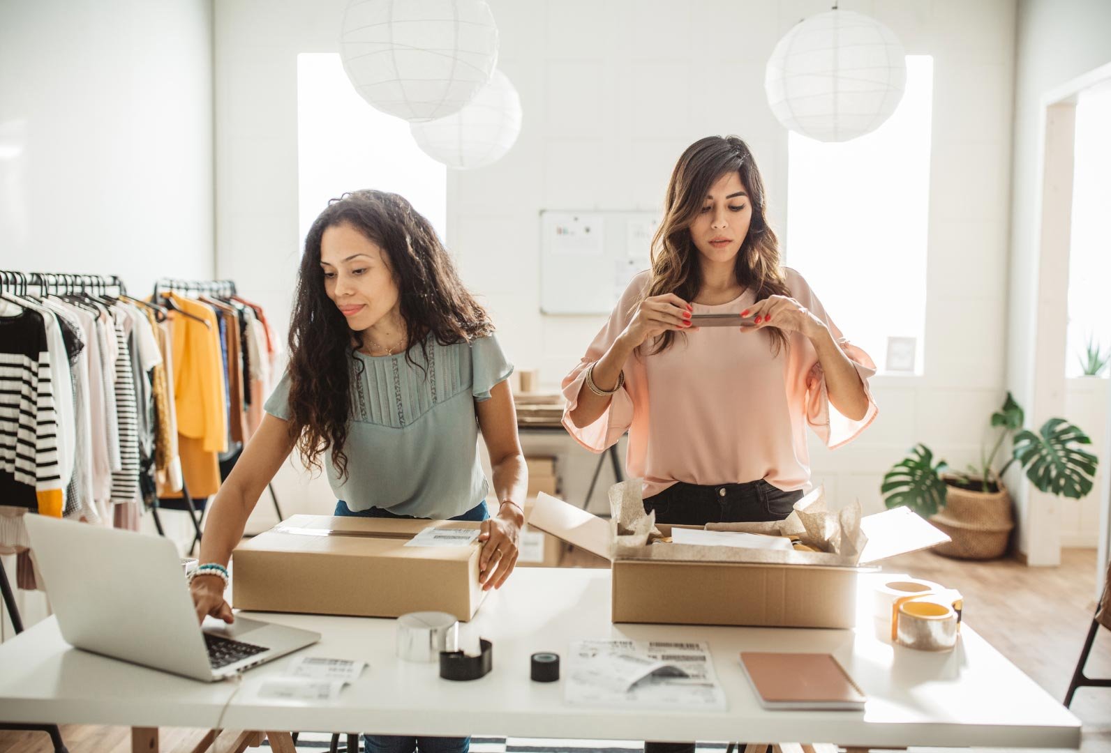 Two women working in a fashion studio, one packing items and the other inspecting a piece of clothing