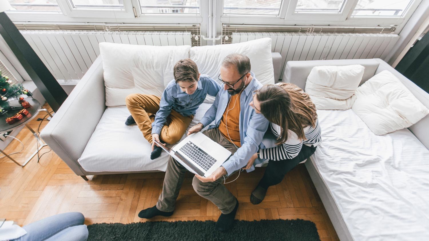 A family of three, including a man, a woman, and a young boy, sitting on a couch looking at a laptop together in a cozy room
